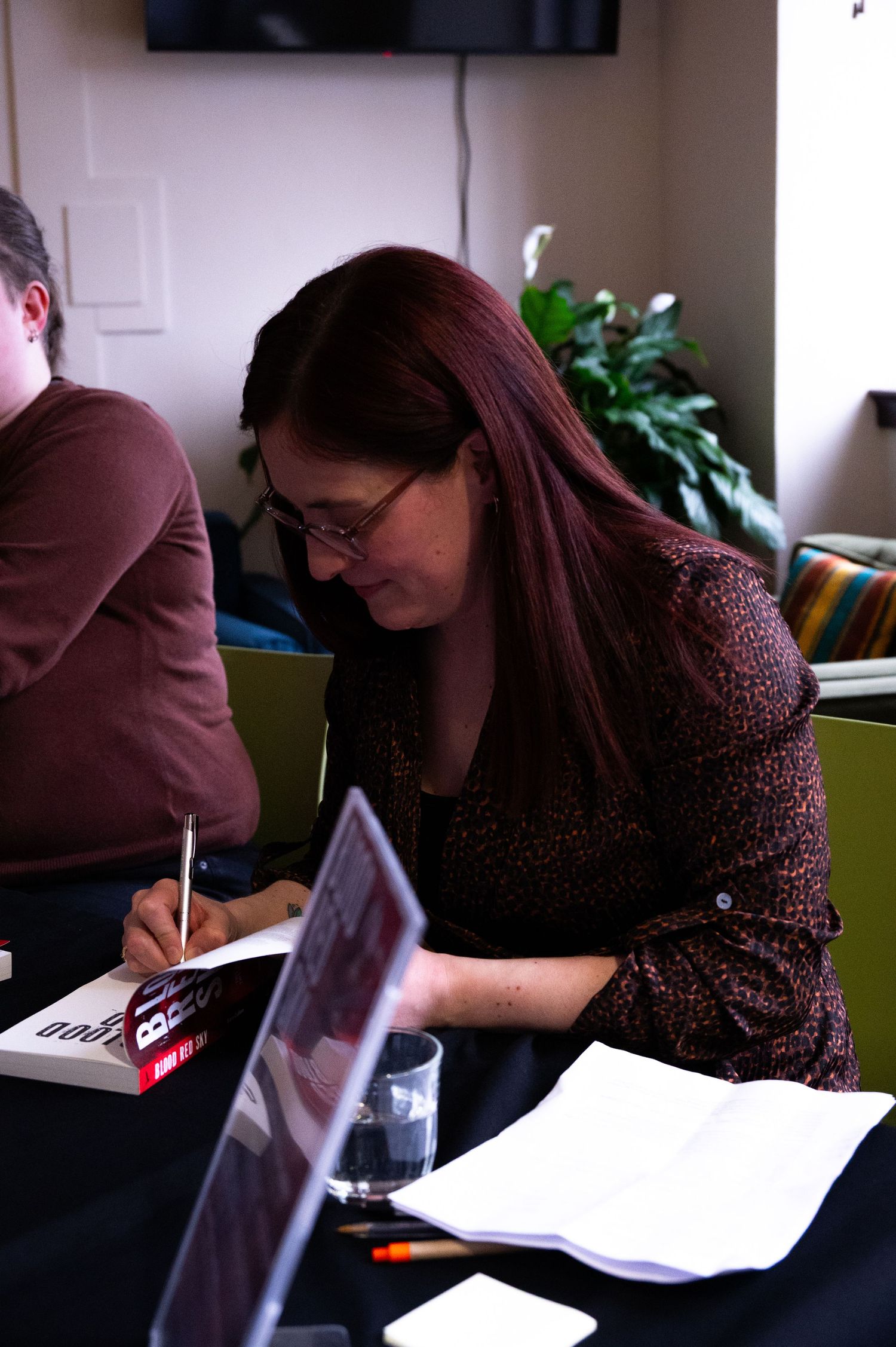 Woman with red hair and glasses signing books at a table during an author signing event.
