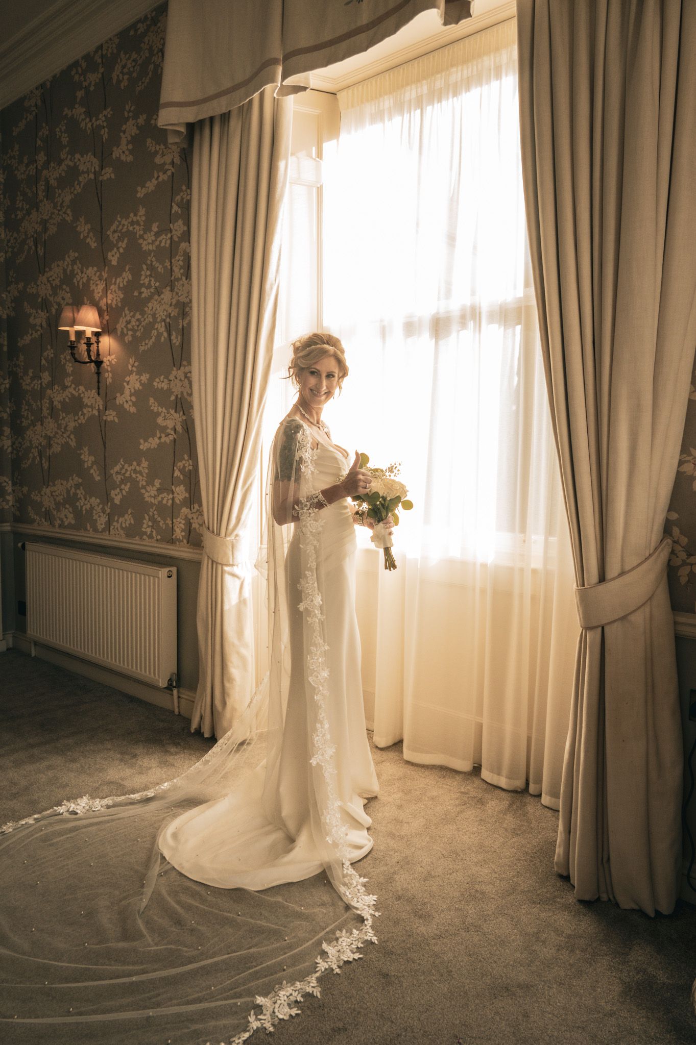 Elegant bride in lace wedding gown with long veil holding bouquet, standing by a sunlit window in ornate room.