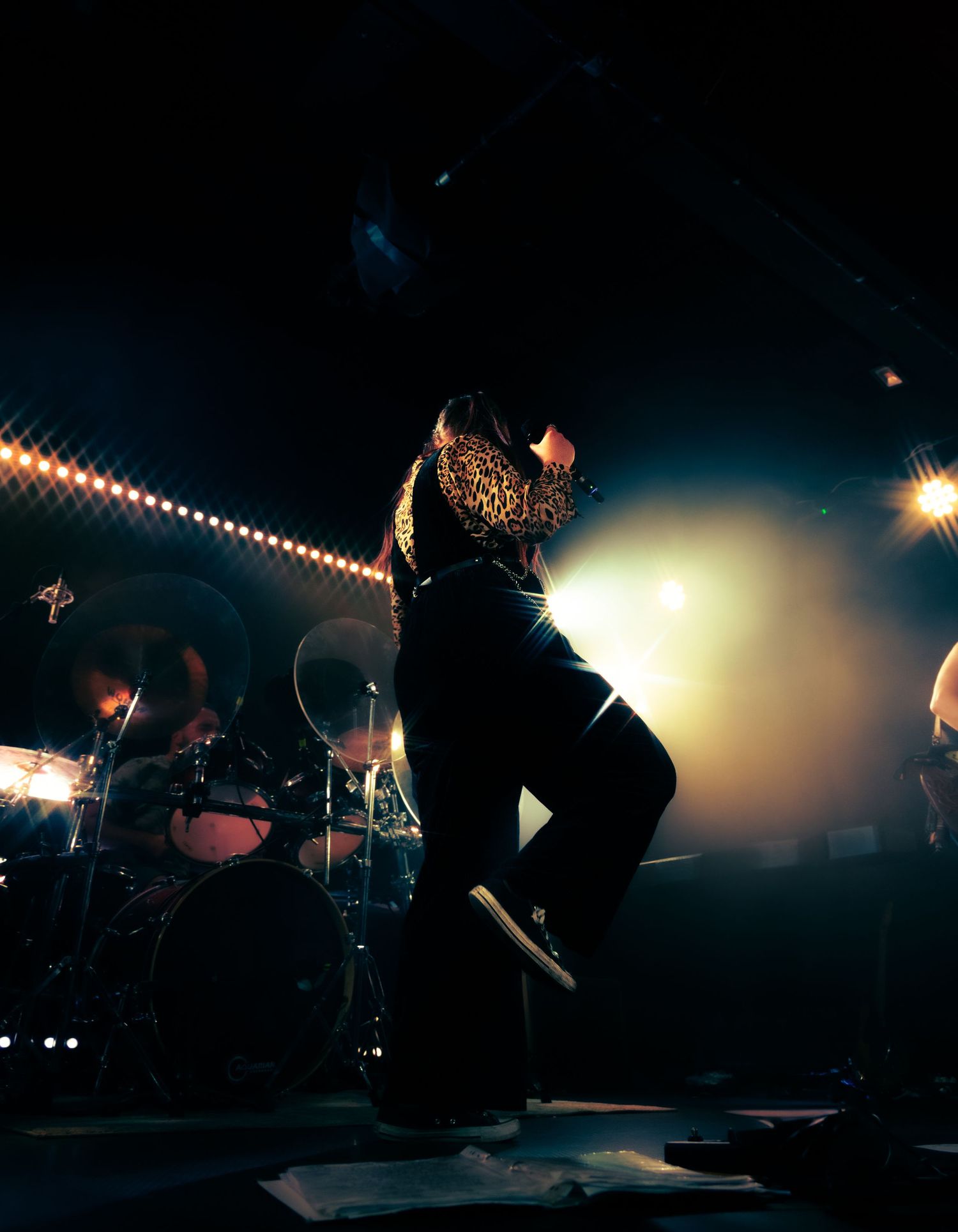 Silhouetted performer jumps on stage while string lights glow overhead during a dramatic concert performance.