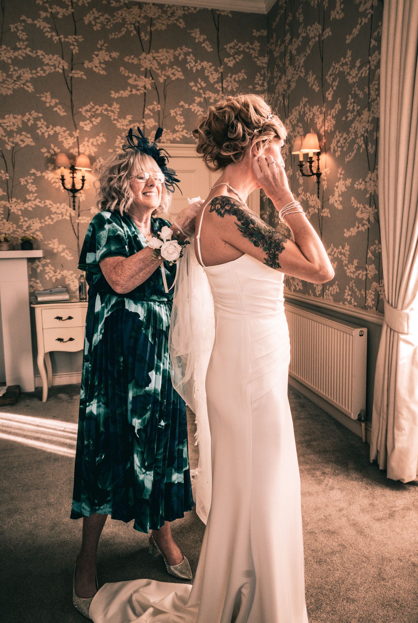 Emotional bride in white gown wiping tears as older woman in teal dress adjusts her veil in elegant room.