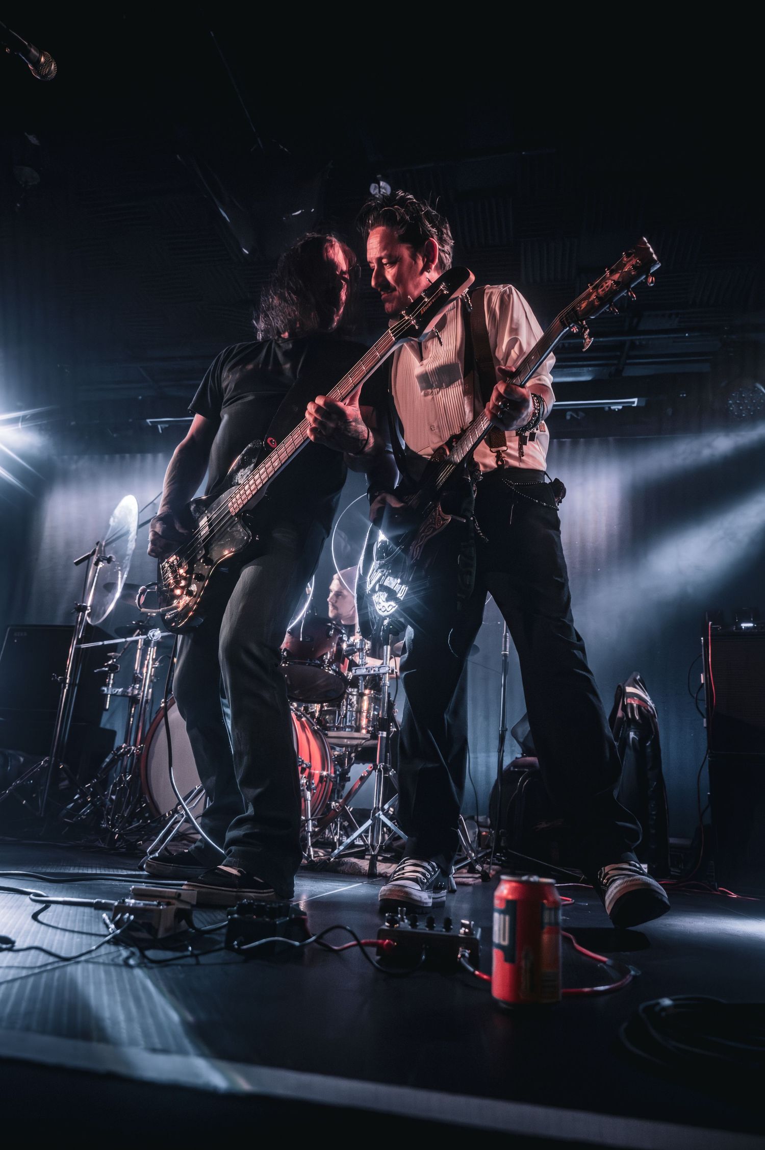 Two guitarists perform back-to-back on a dark concert stage with dramatic lighting and a drummer visible in the background.