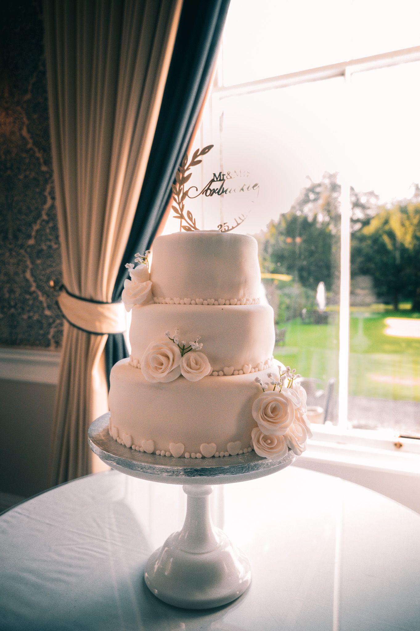 Elegant three-tier white wedding cake with rose decorations and gold topper on a pedestal stand by a window.