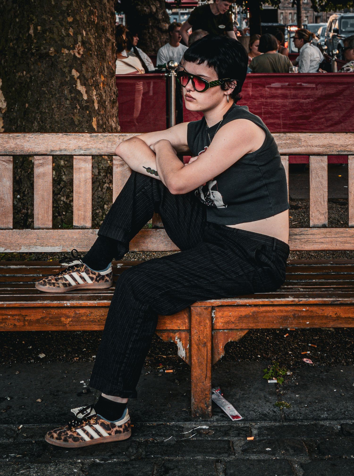 A young woman with short dark hair and red sunglasses sits on a wooden bench wearing a black crop top and striped pants.