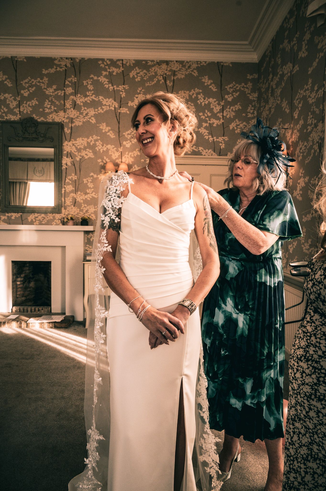 Bride in white gown laughs as older woman in green dress adjusts her lace veil in an elegant wallpapered room.