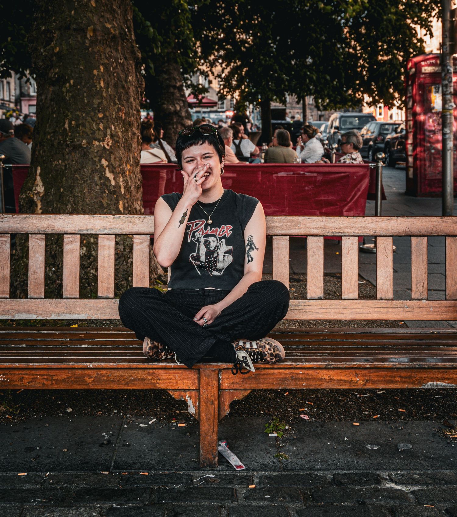 Person sitting cross-legged on a wooden bench outdoors, wearing a black sleeveless shirt and dark pants.