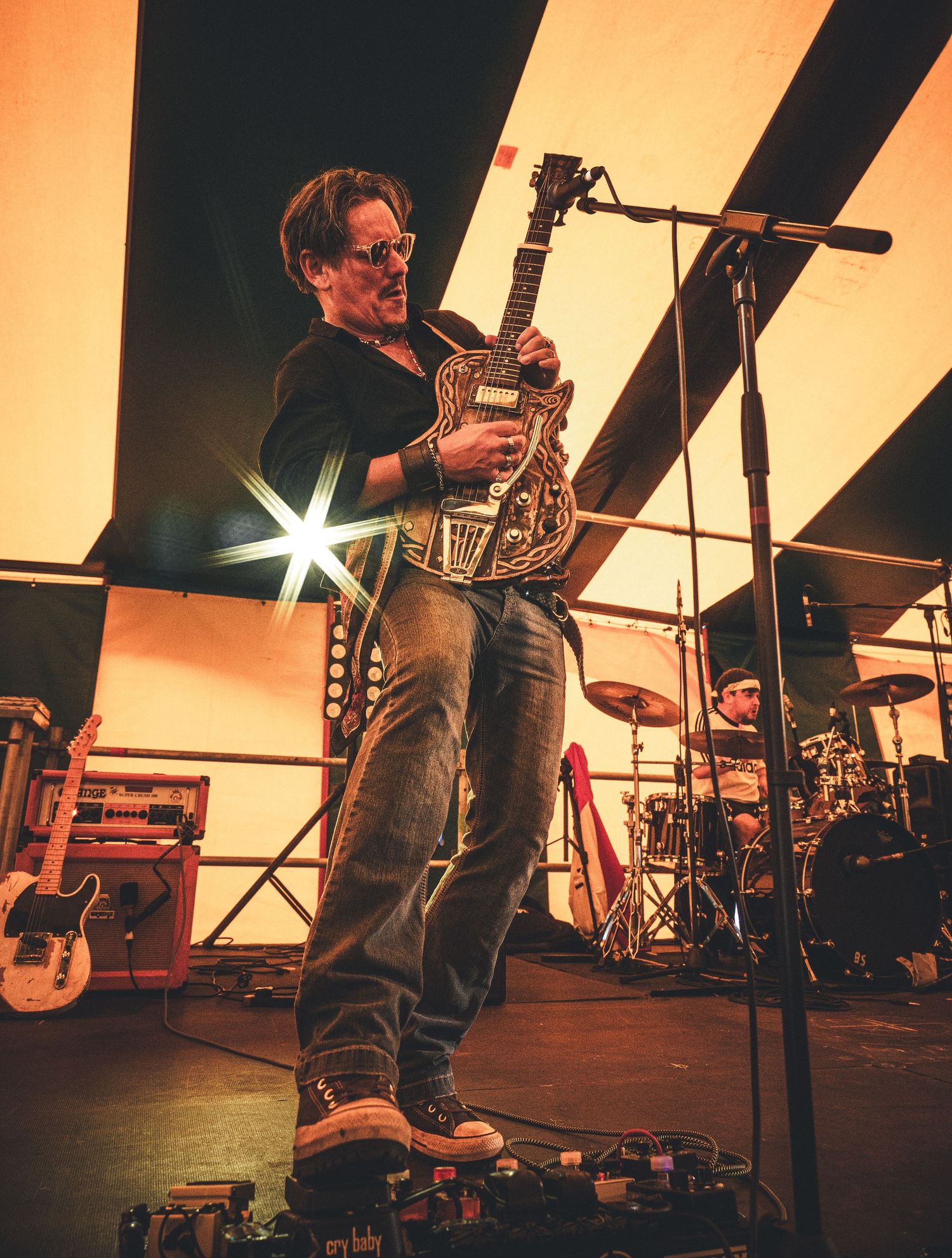 Guitarist performing on stage with ornate guitar under warm lights, drummer visible in background at live music concert.