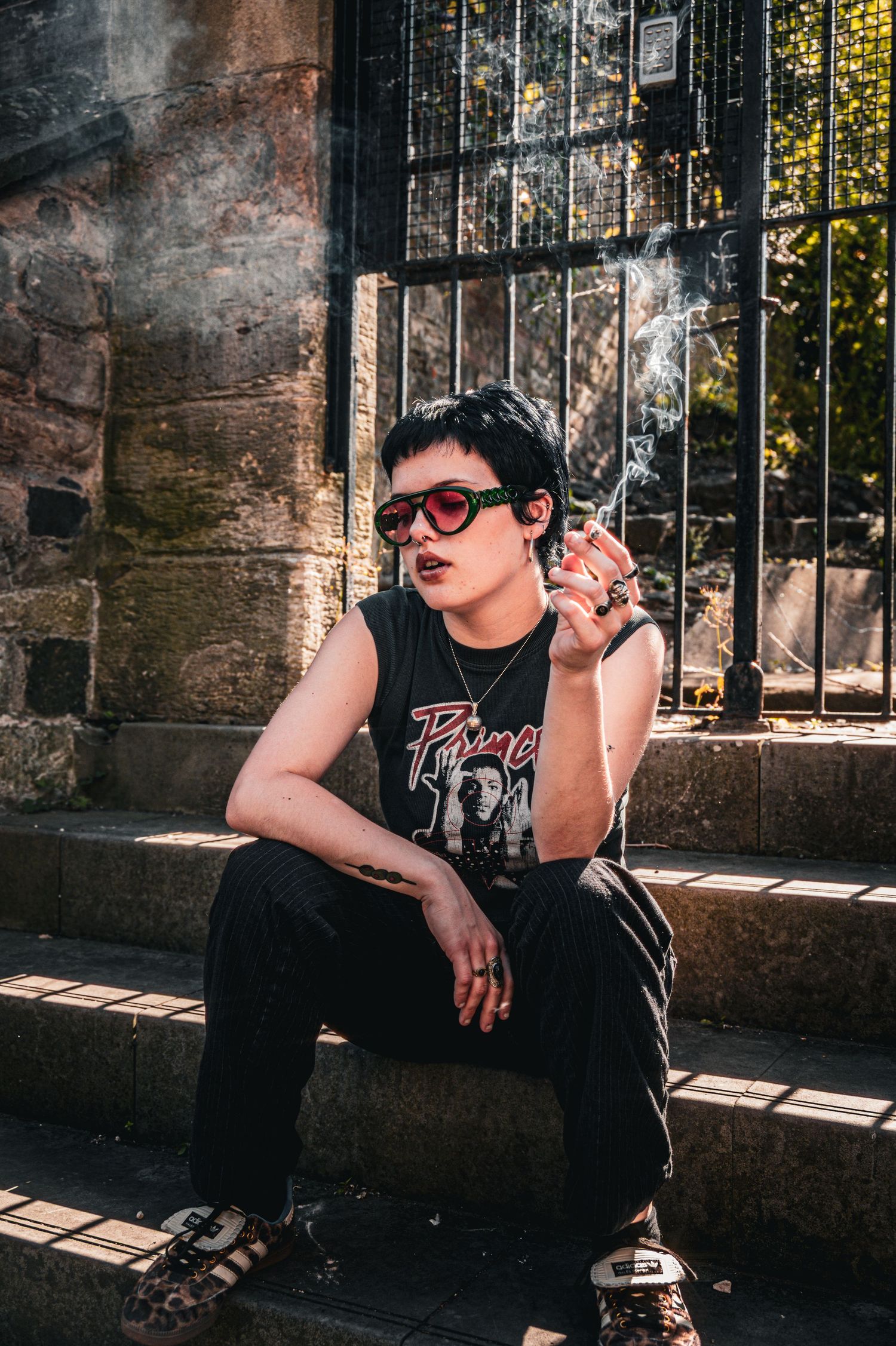 Edgy young woman with short hair and red sunglasses smoking on stone steps in black sleeveless band tee.