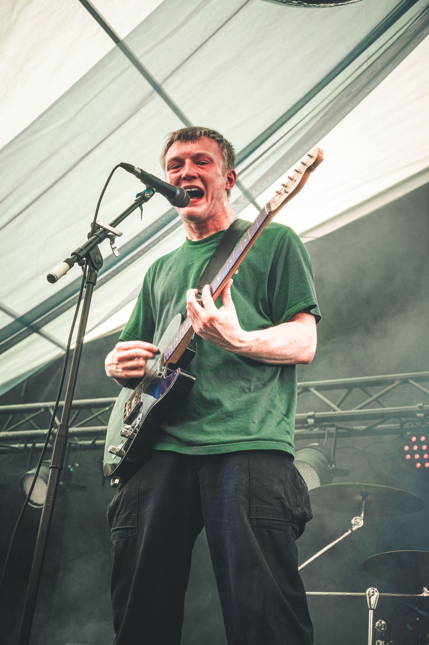 Energetic guitarist in green t-shirt singing into microphone while playing electric guitar on stage.