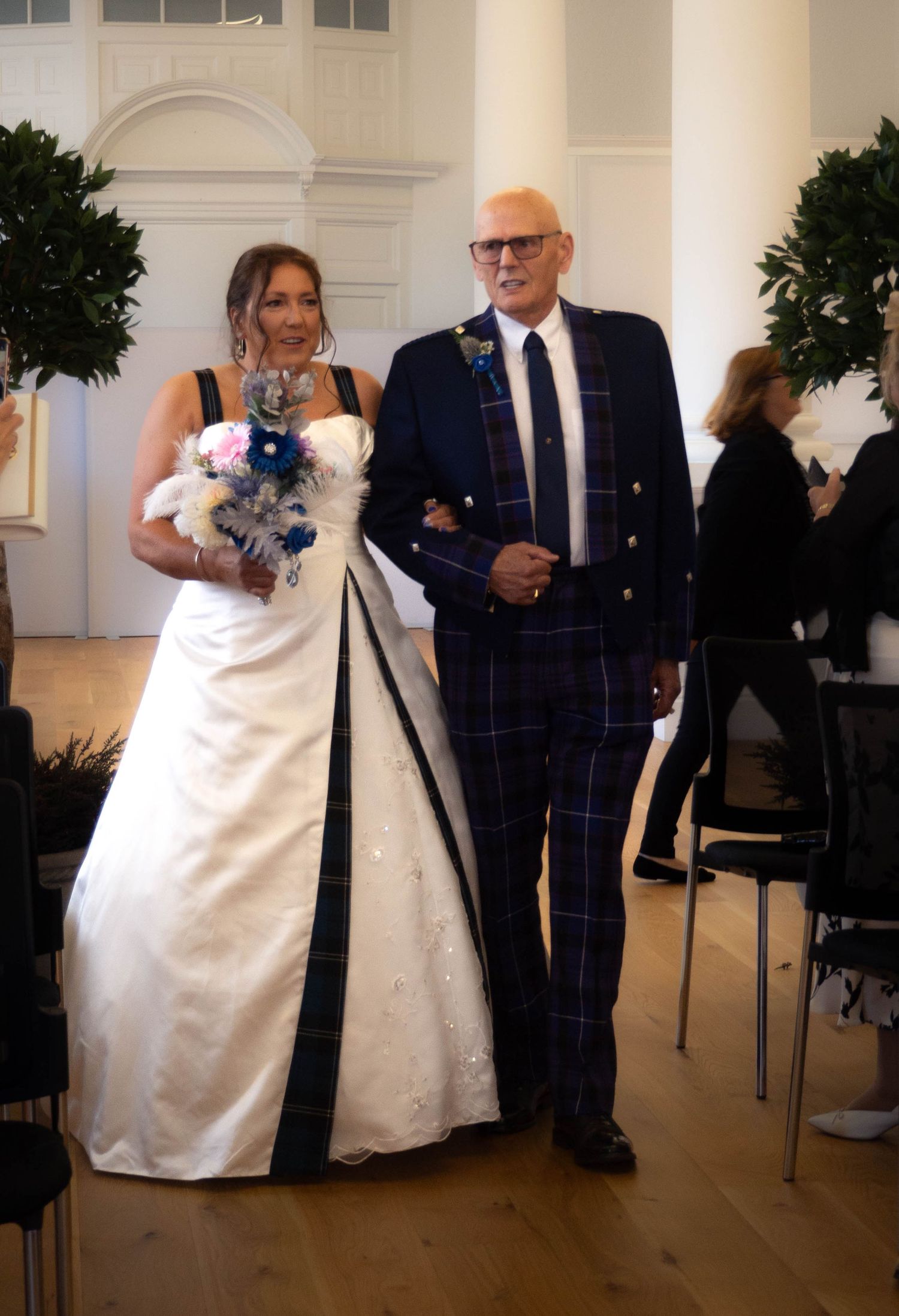 Bride in white gown with tartan trim walks with man in blue tartan suit at indoor wedding ceremony.