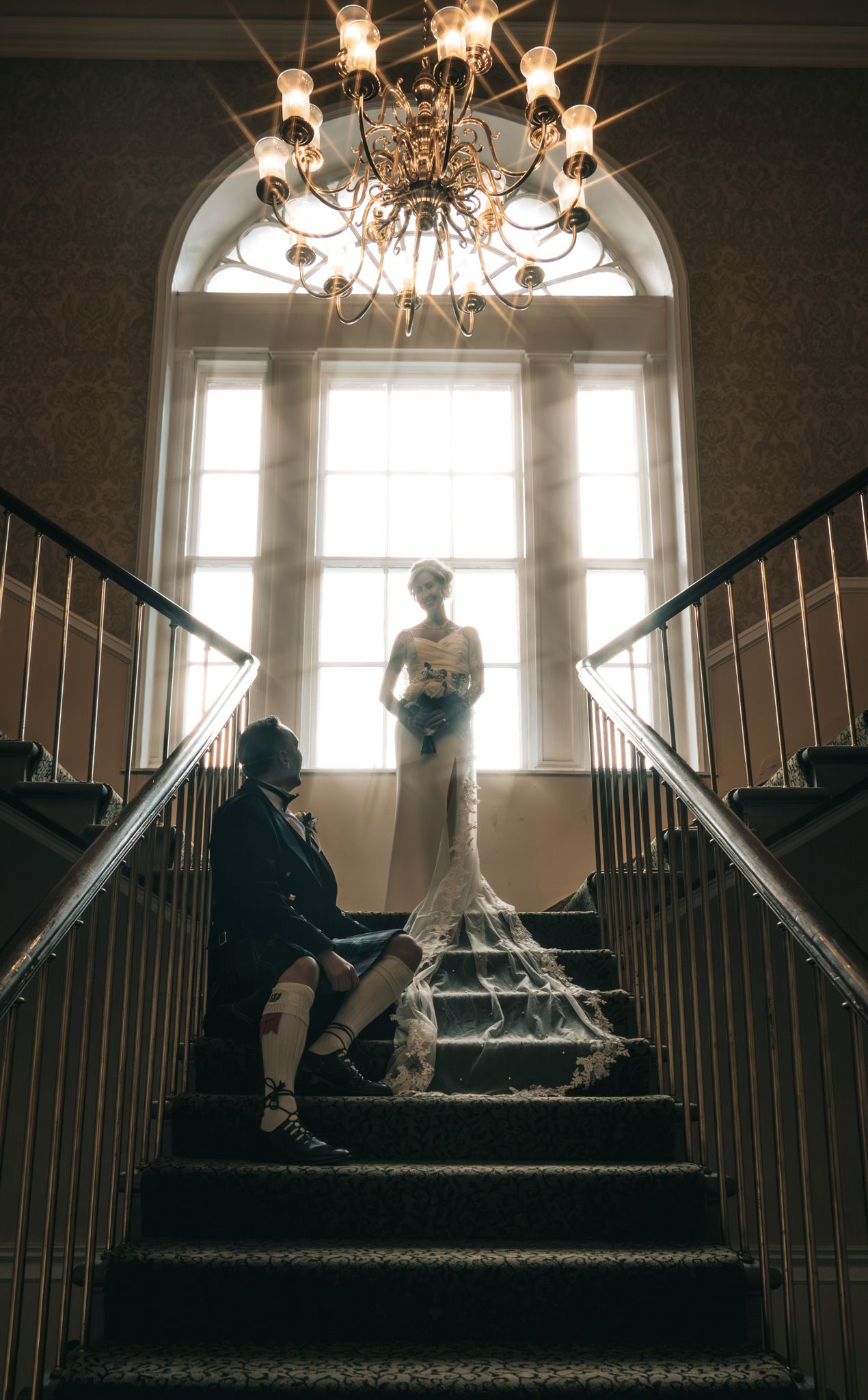 Bride in elegant gown stands atop grand staircase as groom sits below, lit by chandelier and arched window.
