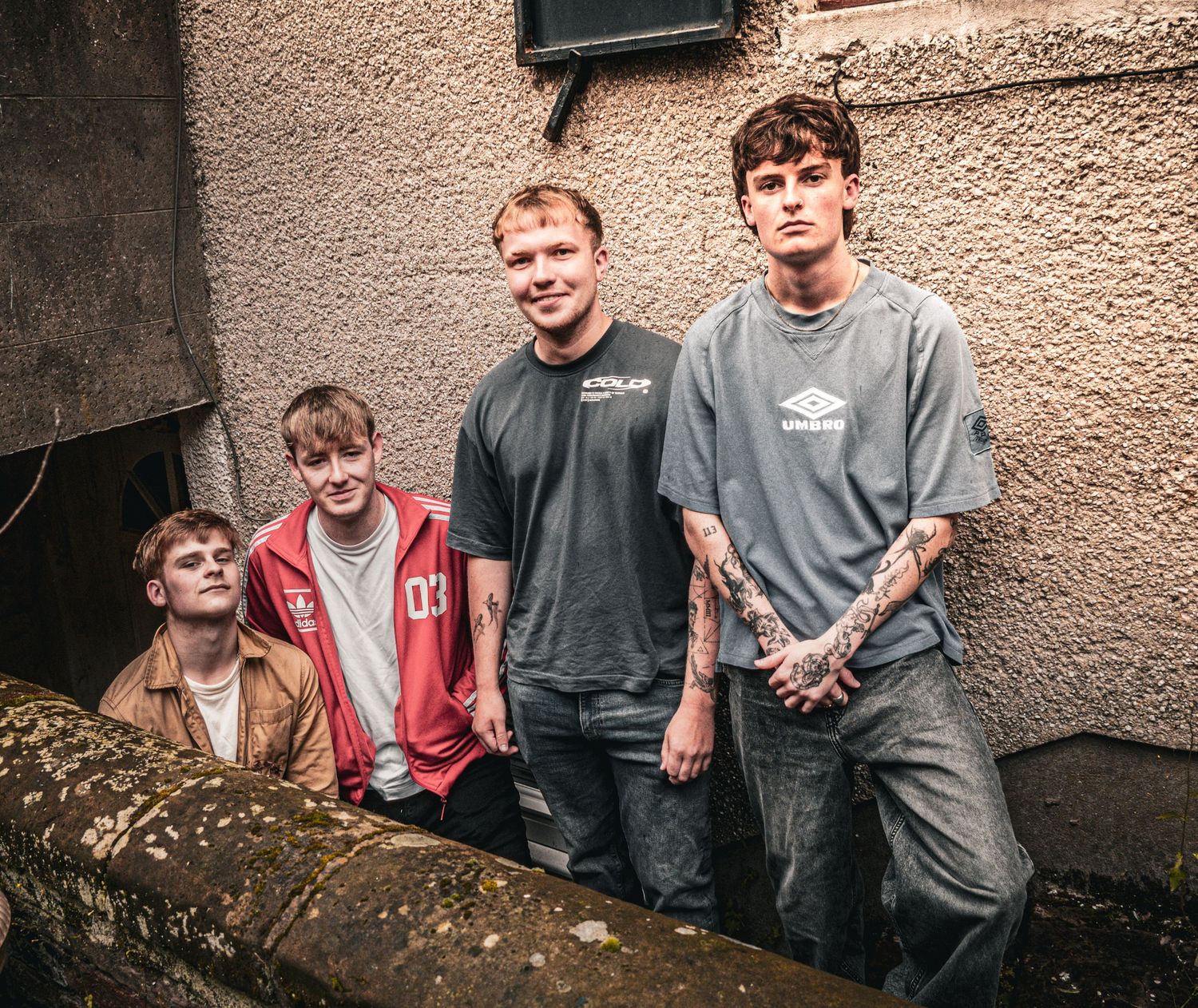 Four young men pose outdoors against a stone wall, wearing casual streetwear including a red hoodie and grey tees.