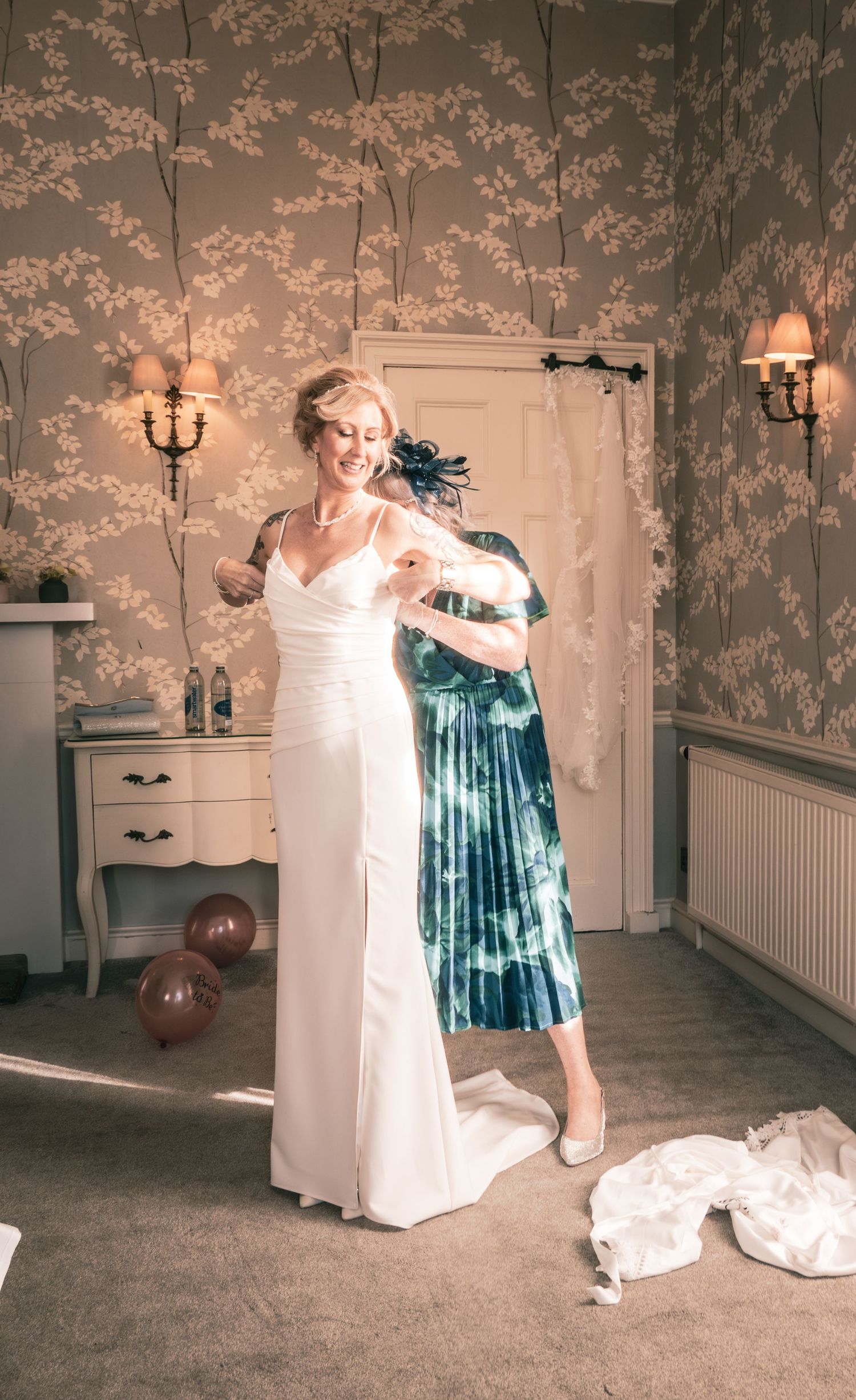 Bride in white gown being helped by bridesmaid in teal dress in elegant floral wallpapered bridal suite.