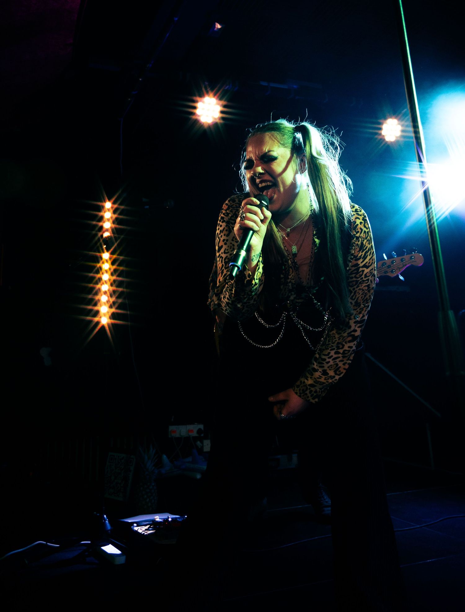 Rock performer in leopard print jacket sings passionately on darkly lit stage with dramatic overhead lighting effects.
