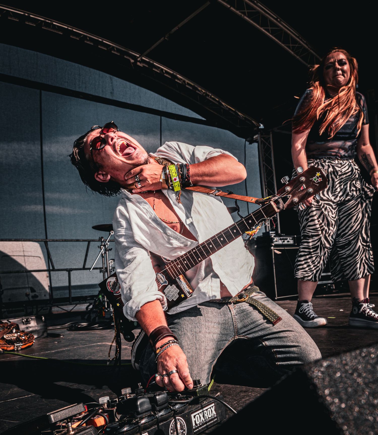 A guitarist dramatically kneels on stage while a female guitarist performs behind him outdoors.