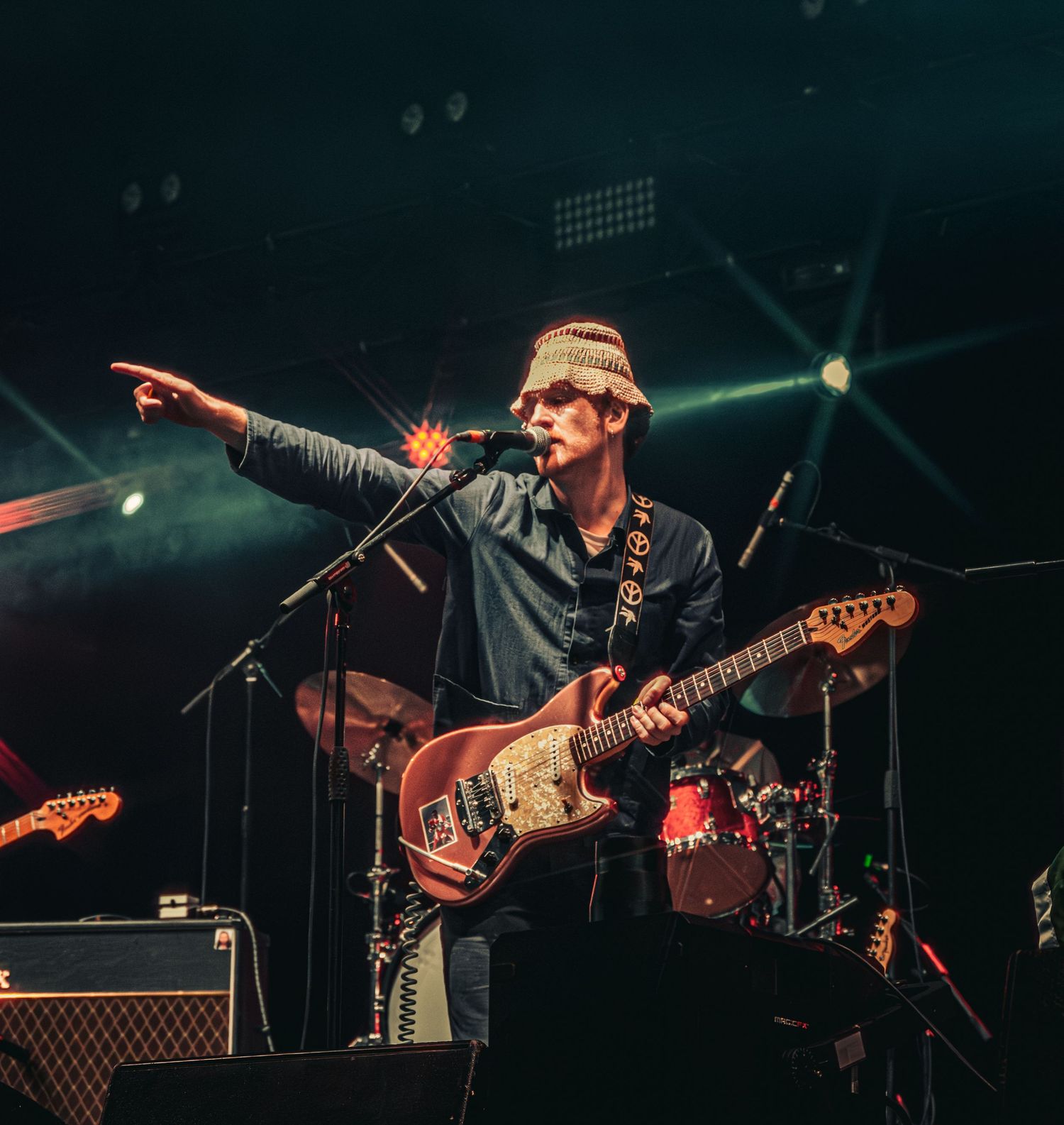 Guitarist in a beanie hat points to the crowd while singing on a dramatic stage with colorful lights.