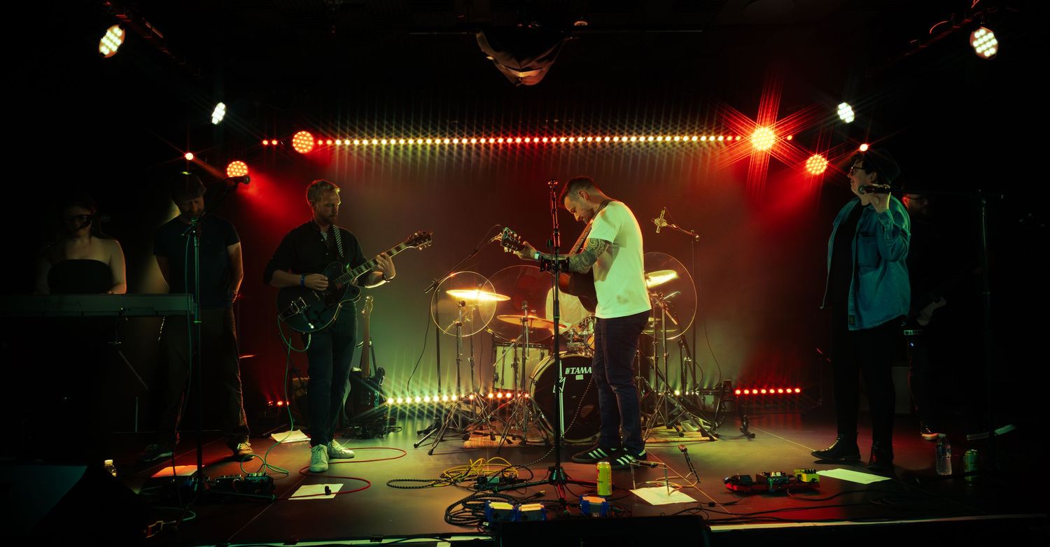 Band performs on dimly lit stage with red and green lighting effects behind illuminated LED strip light backdrop.