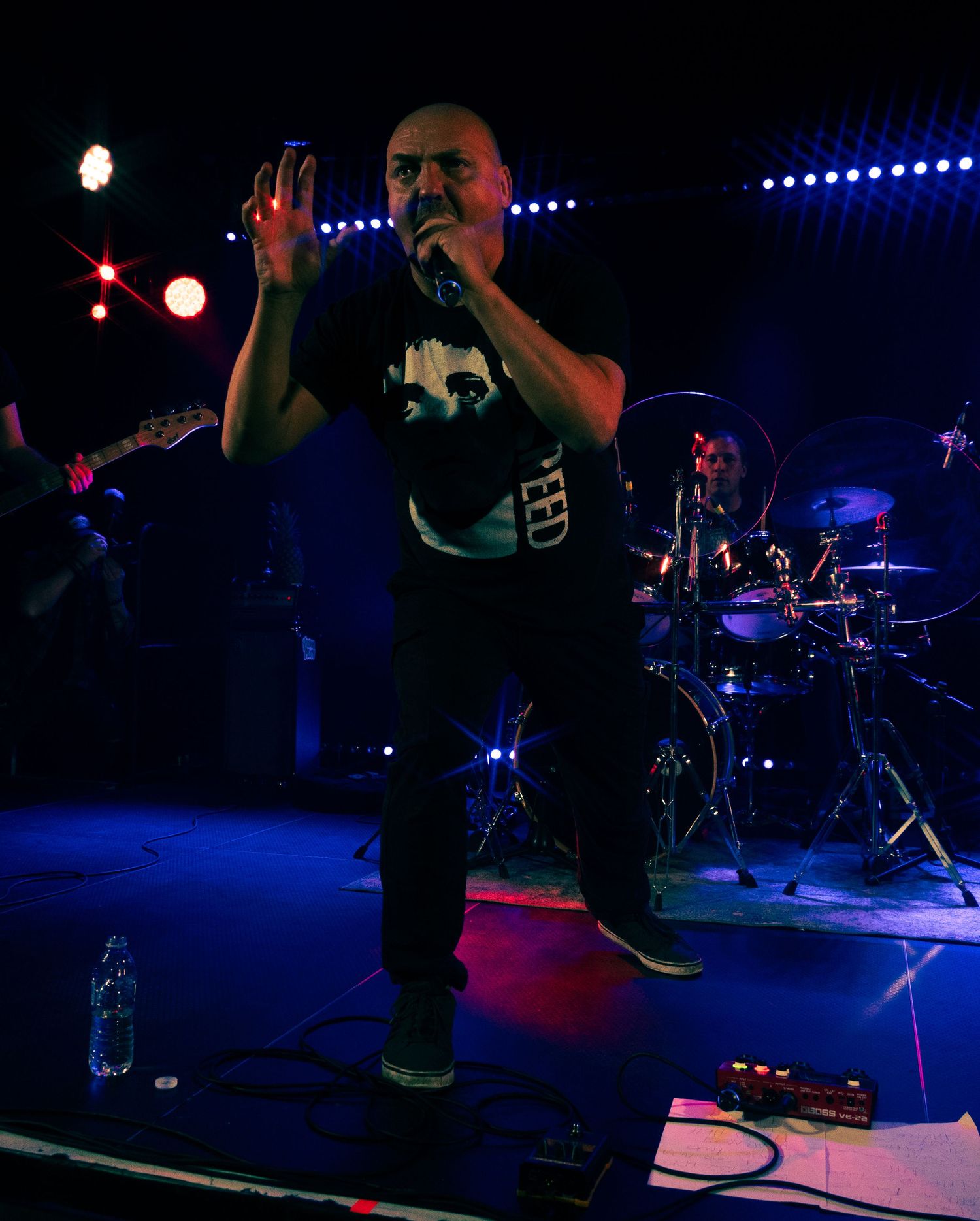 A performer in a dark shirt energetically sings on stage with blue lighting and a drum set visible in the background.