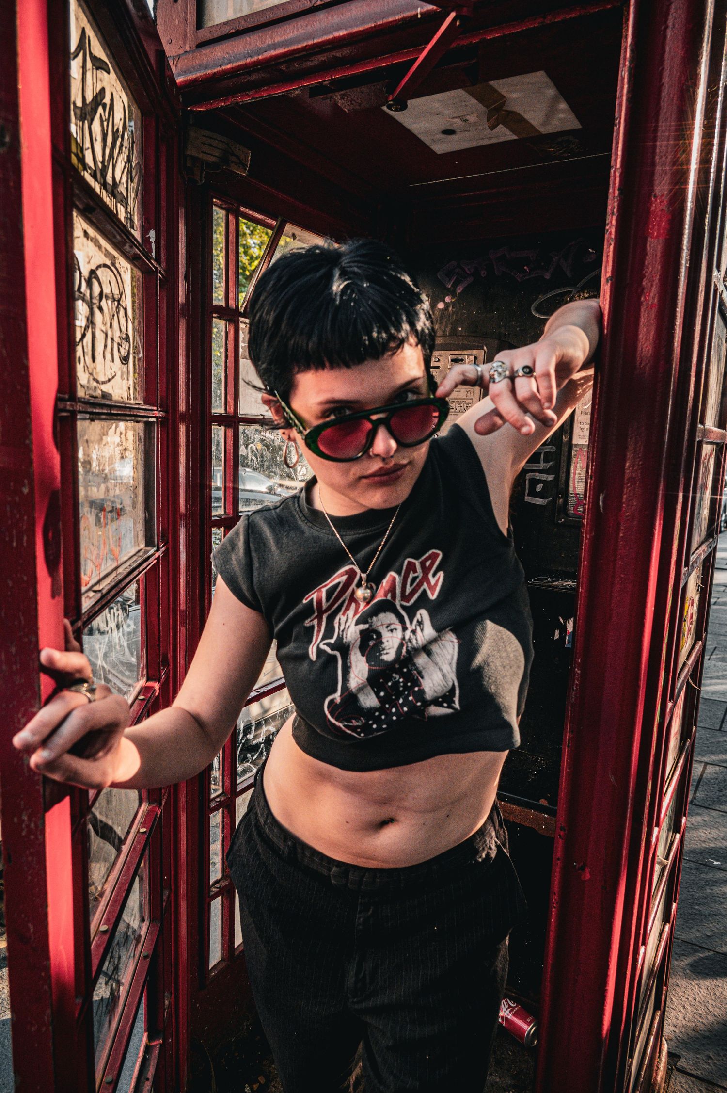 Person in a Prince crop tee and red sunglasses posing inside a graffiti-covered red phone booth.