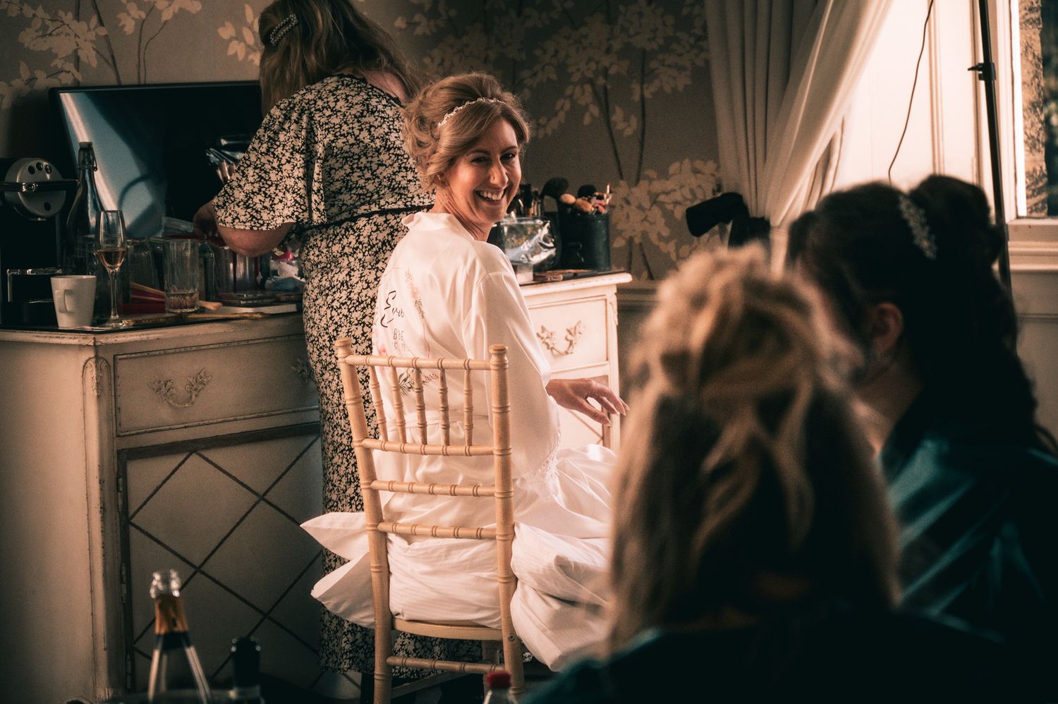 Bride in white robe laughing with bridesmaids during wedding preparations in an elegant dressing room.