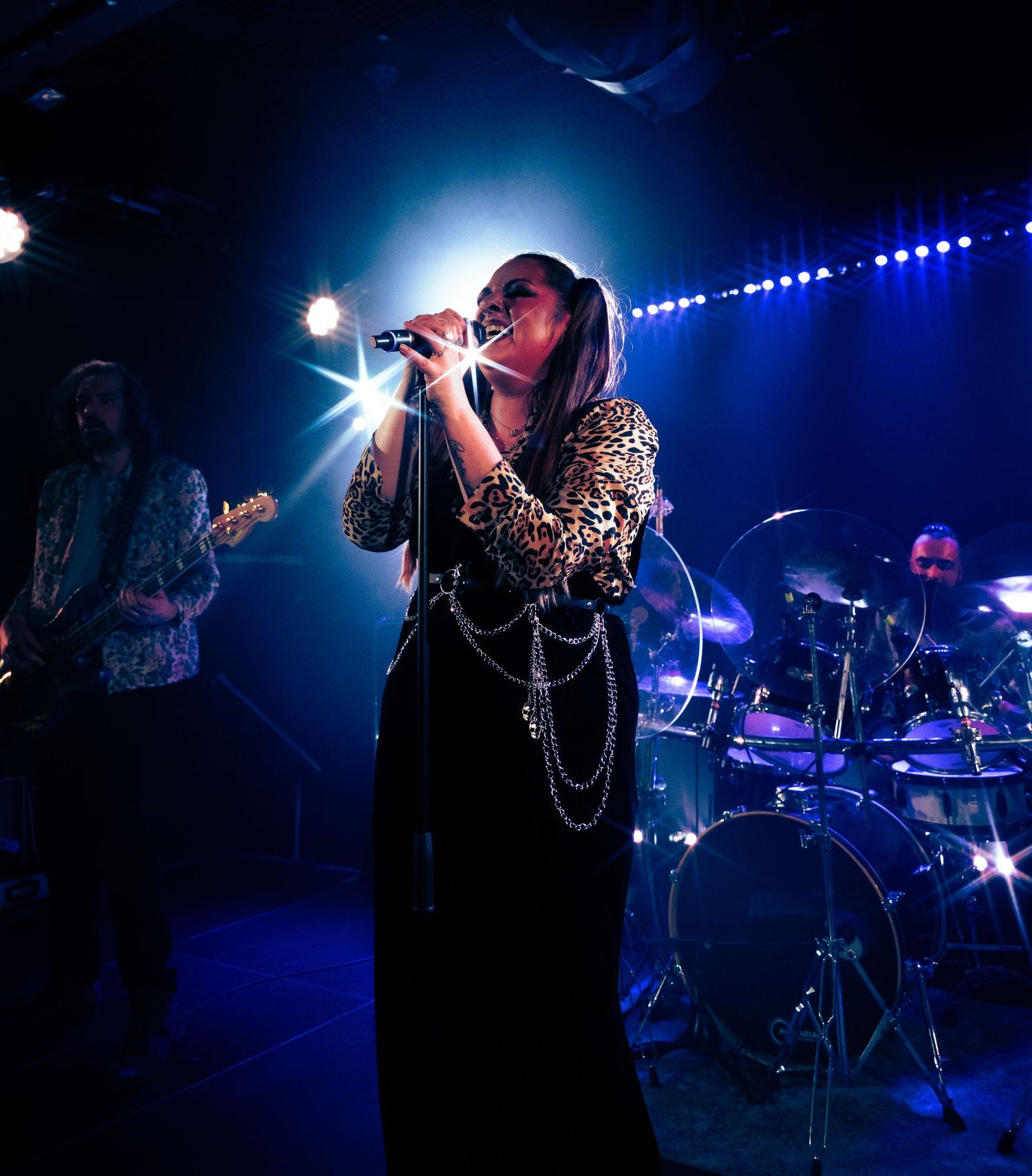 A singer performs on stage in a long black dress and chains with dramatic blue lighting and band members visible in background.