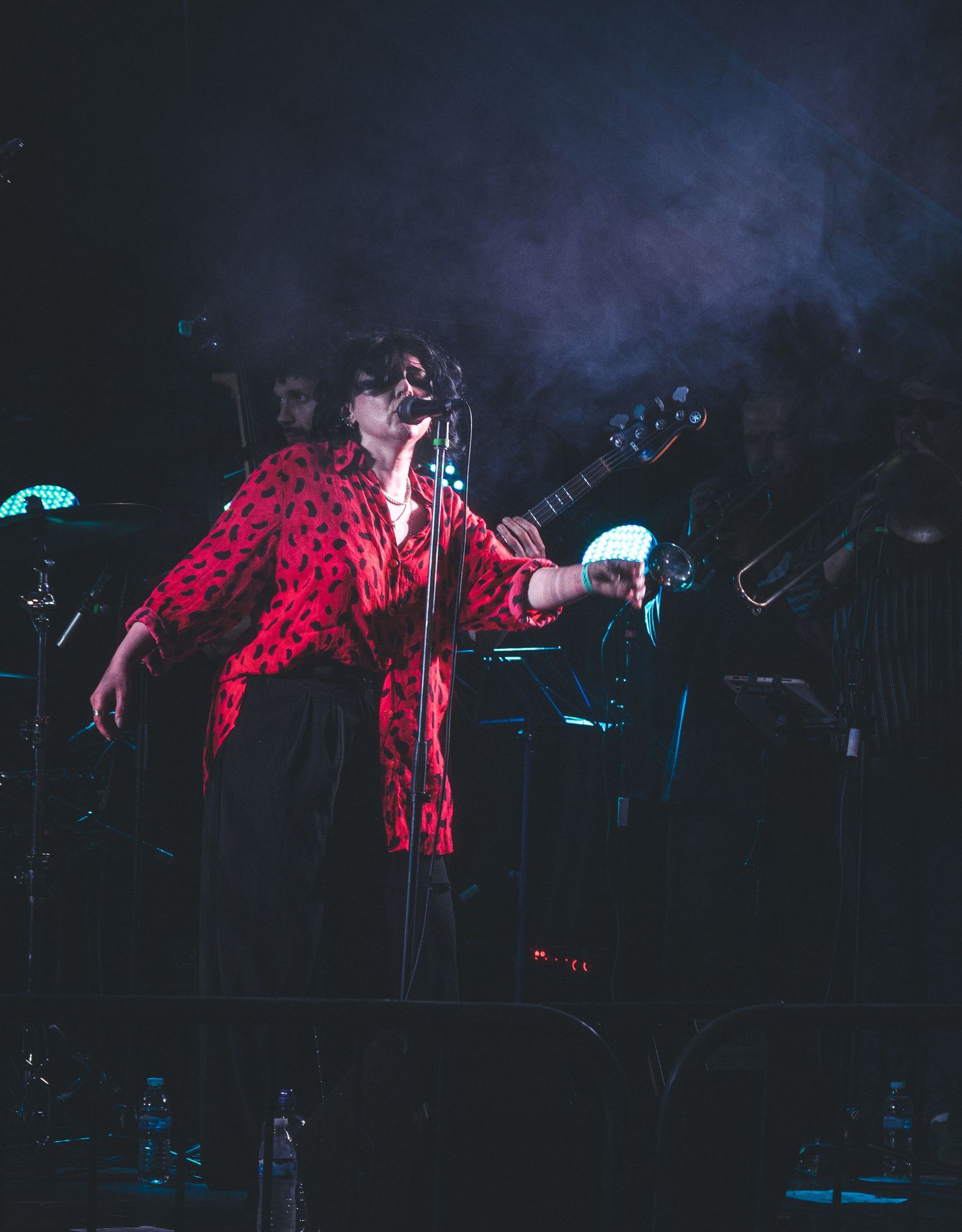 A performer in a red polka dot shirt sings passionately on a dimly lit stage with band members visible behind.