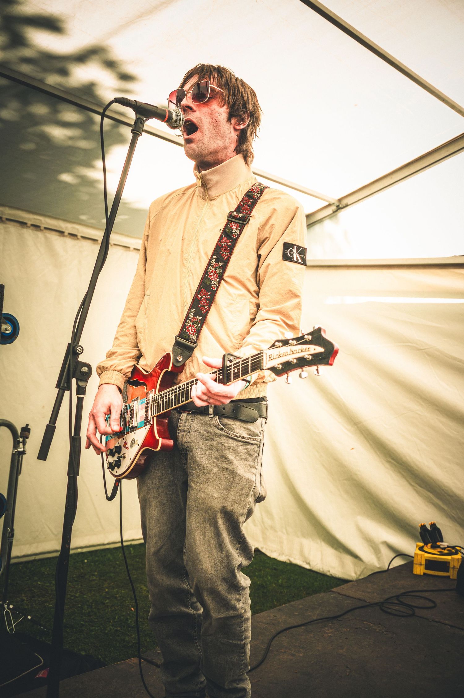 Guitarist singer performing on stage wearing sunglasses and beige shirt playing red electric guitar at outdoor festival.