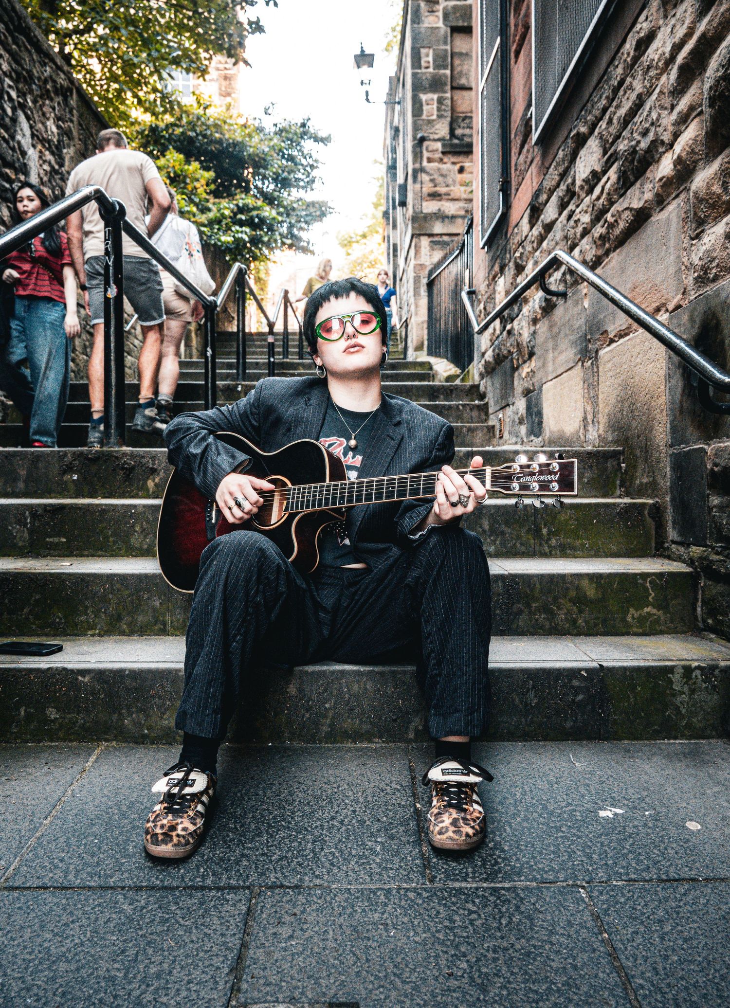 Young guitarist playing acoustic guitar on city steps wearing red sunglasses and black outfit.