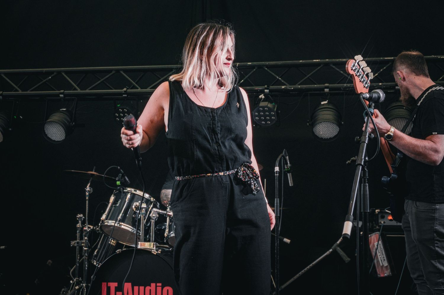 Female vocalist in black jumpsuit performing on stage with guitarist in background.