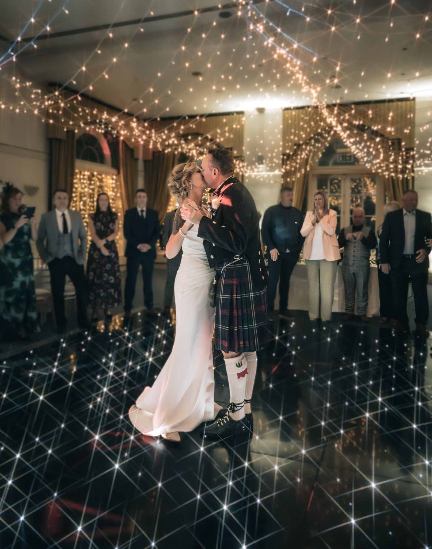 Couple sharing their first dance on a sparkling dance floor under fairy lights in a kilt.