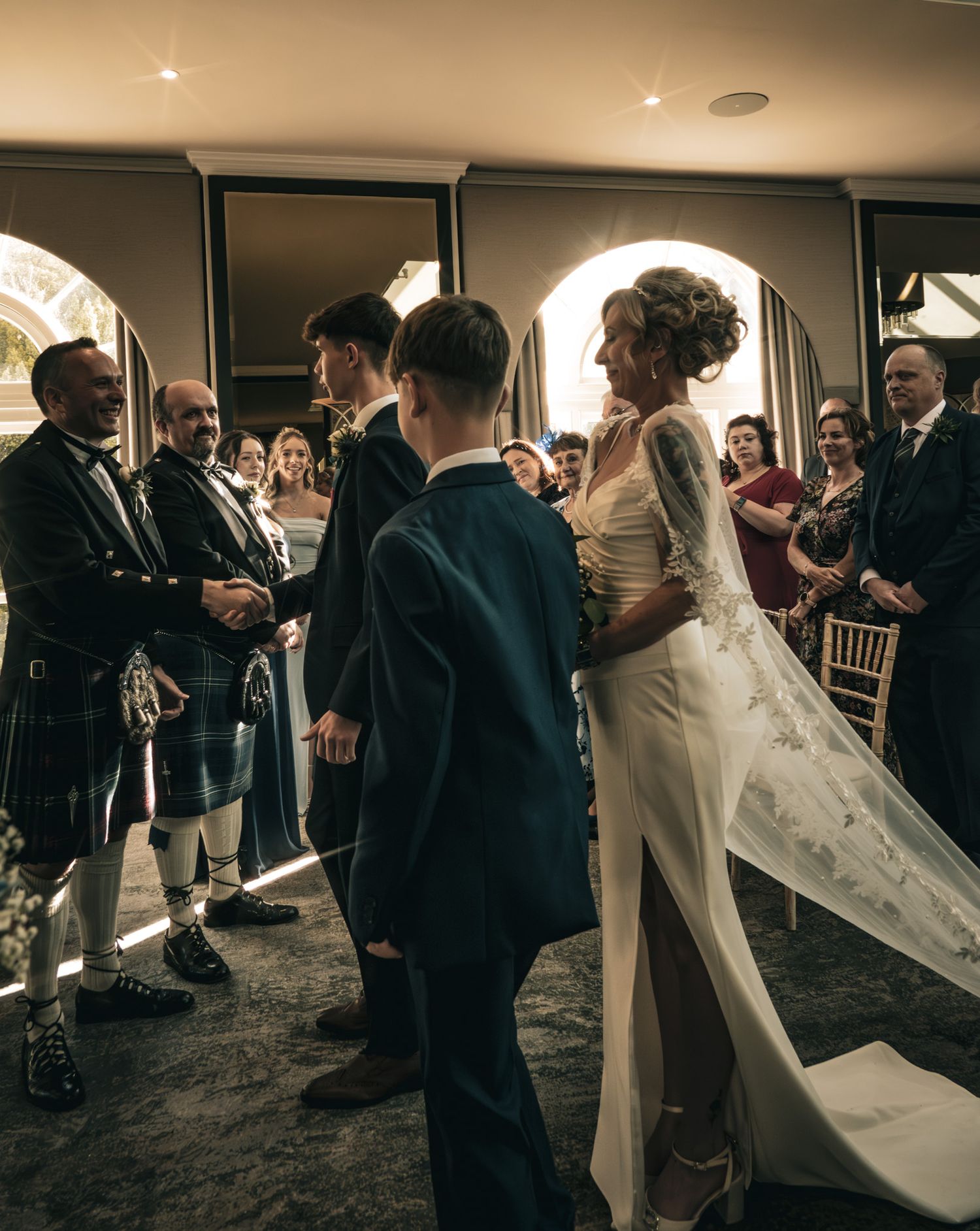Bride in white gown with train walks down aisle flanked by guests in kilts at elegant wedding ceremony.