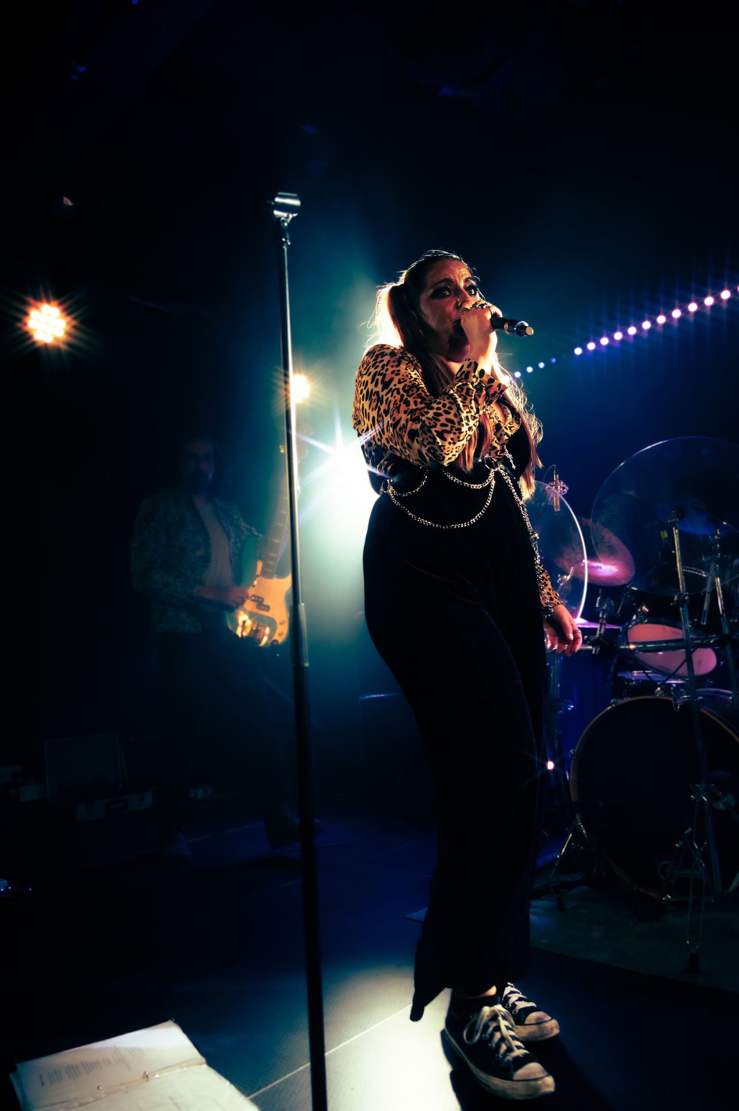 Singer performing on stage in dark lighting wearing black outfit against blue lights and drum kit backdrop.