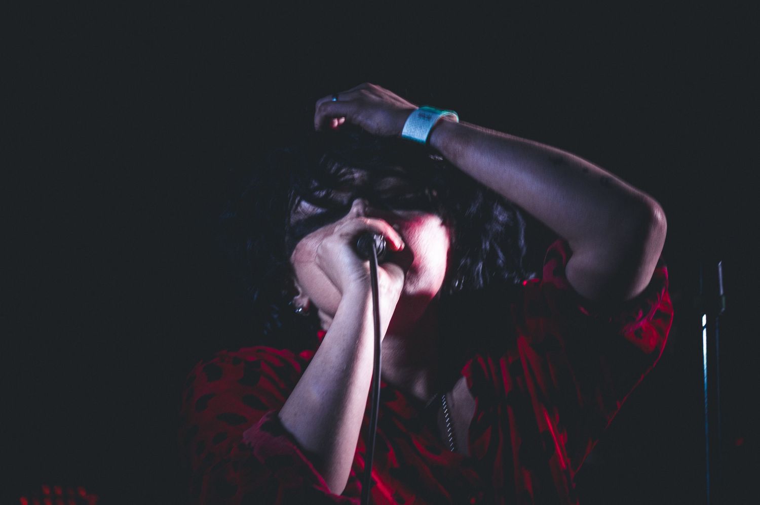 A performer in a red shirt holds a microphone, head bowed with hands raised, under dramatic stage lighting.