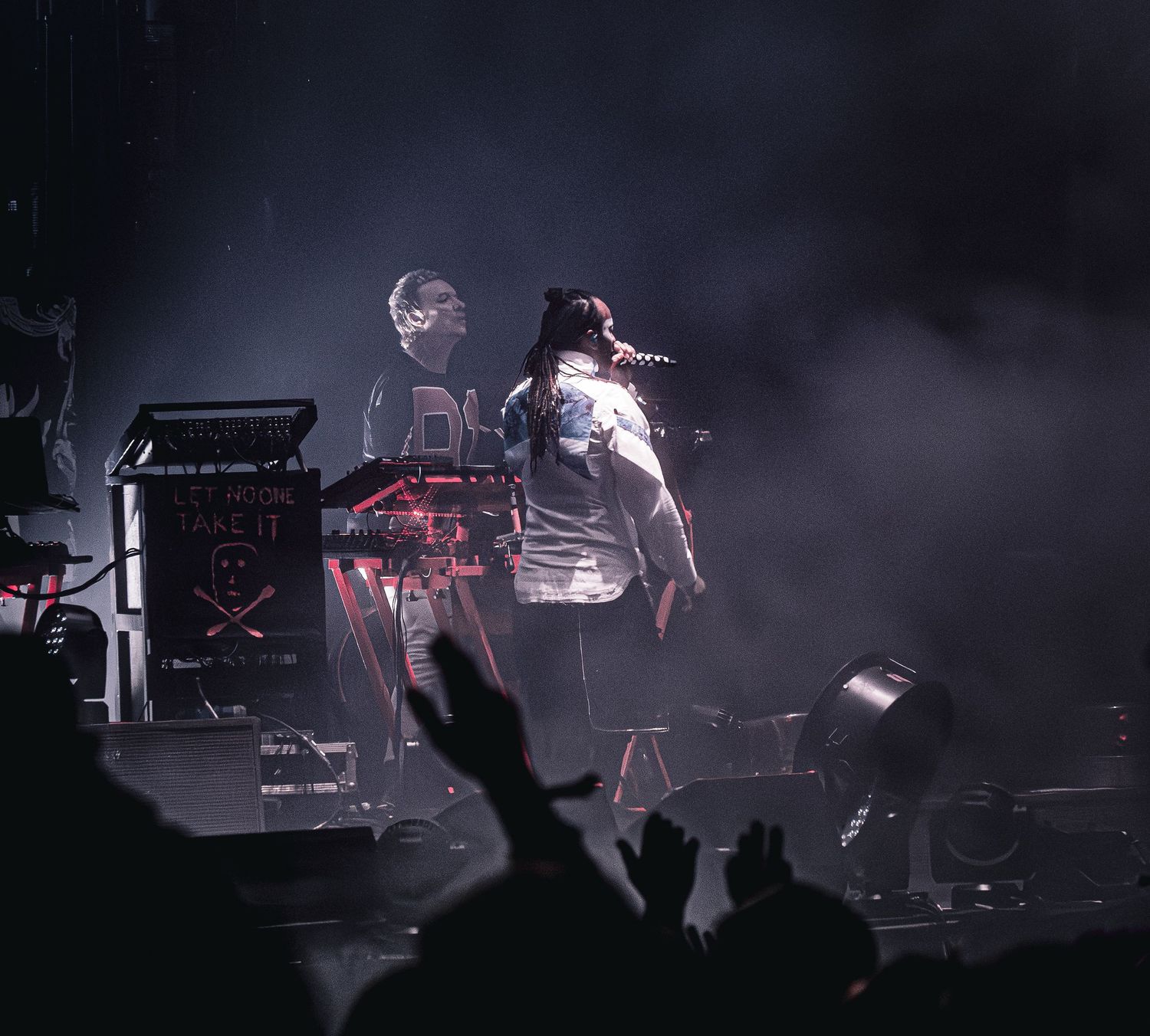 Two performers on a smoky stage, one at keyboards and one singing into a mic, with hands raised crowd.