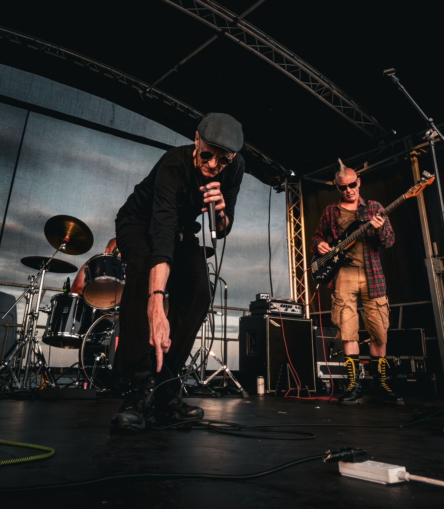 Rock band performing on outdoor stage, vocalist leaning forward with microphone and guitarist in background.