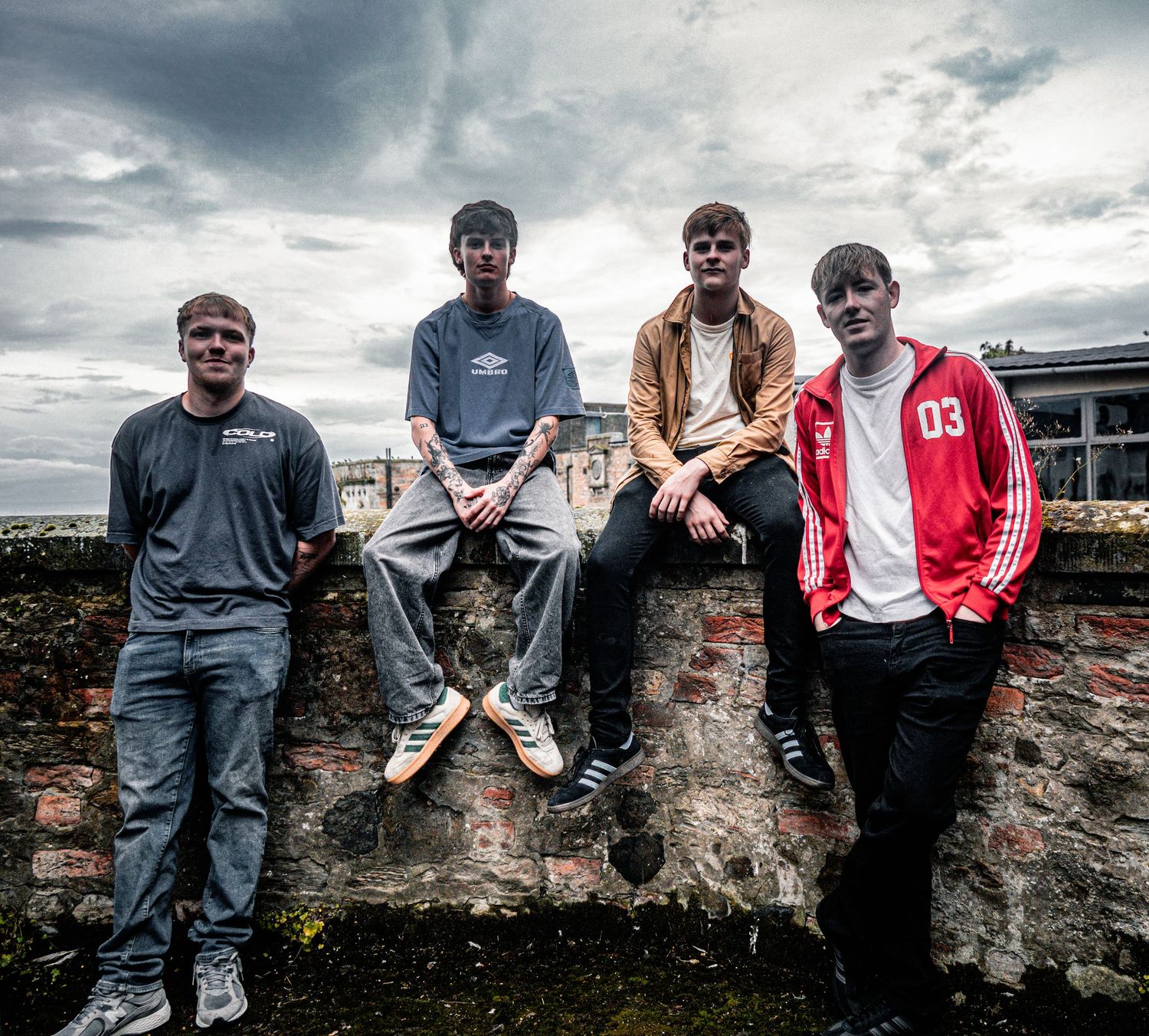 Four young men pose outdoors on a stone wall under a dramatic cloudy sky, one wearing a red Umbro jacket.