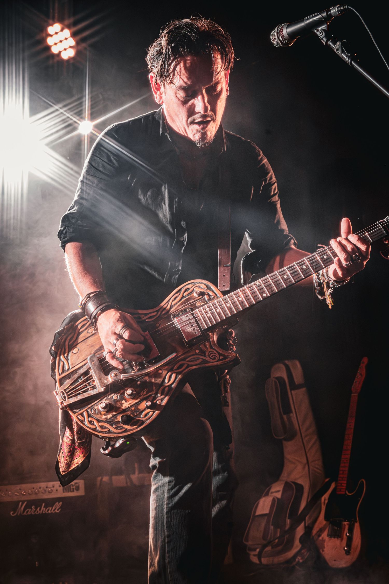 Male guitarist performing on stage with a decorative guitar under dramatic stage lighting and fog effects.