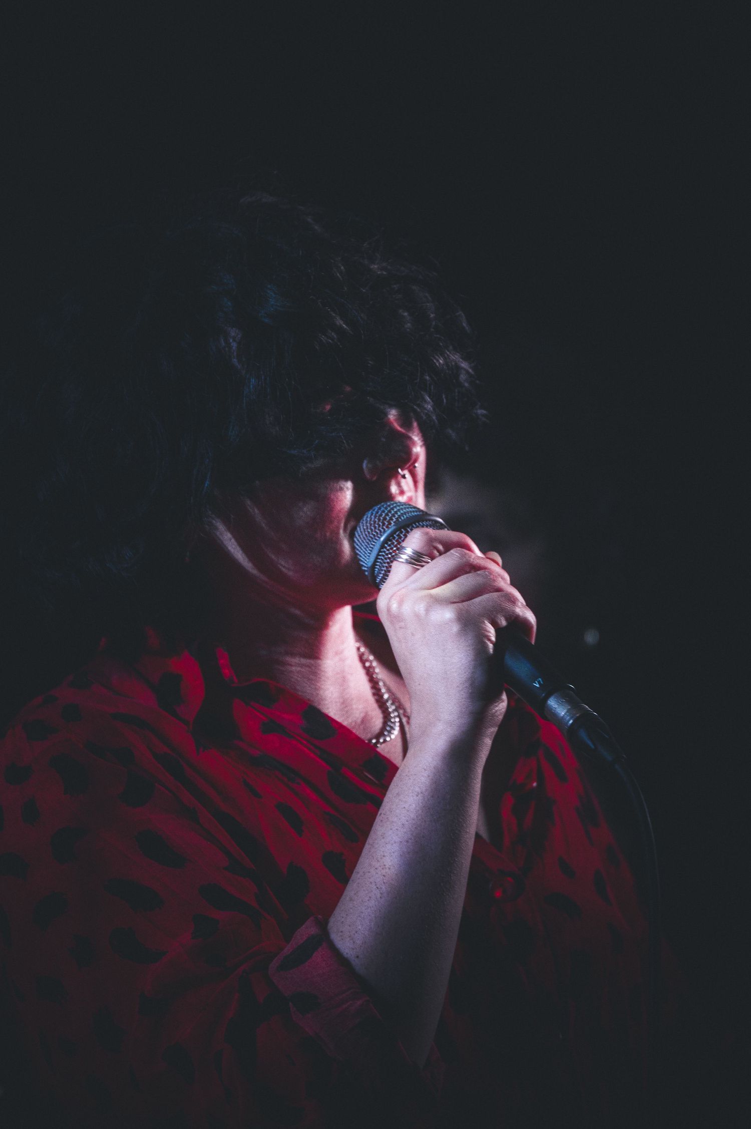 A singer in a red floral top performs passionately on stage, holding a microphone close in dramatic low lighting.