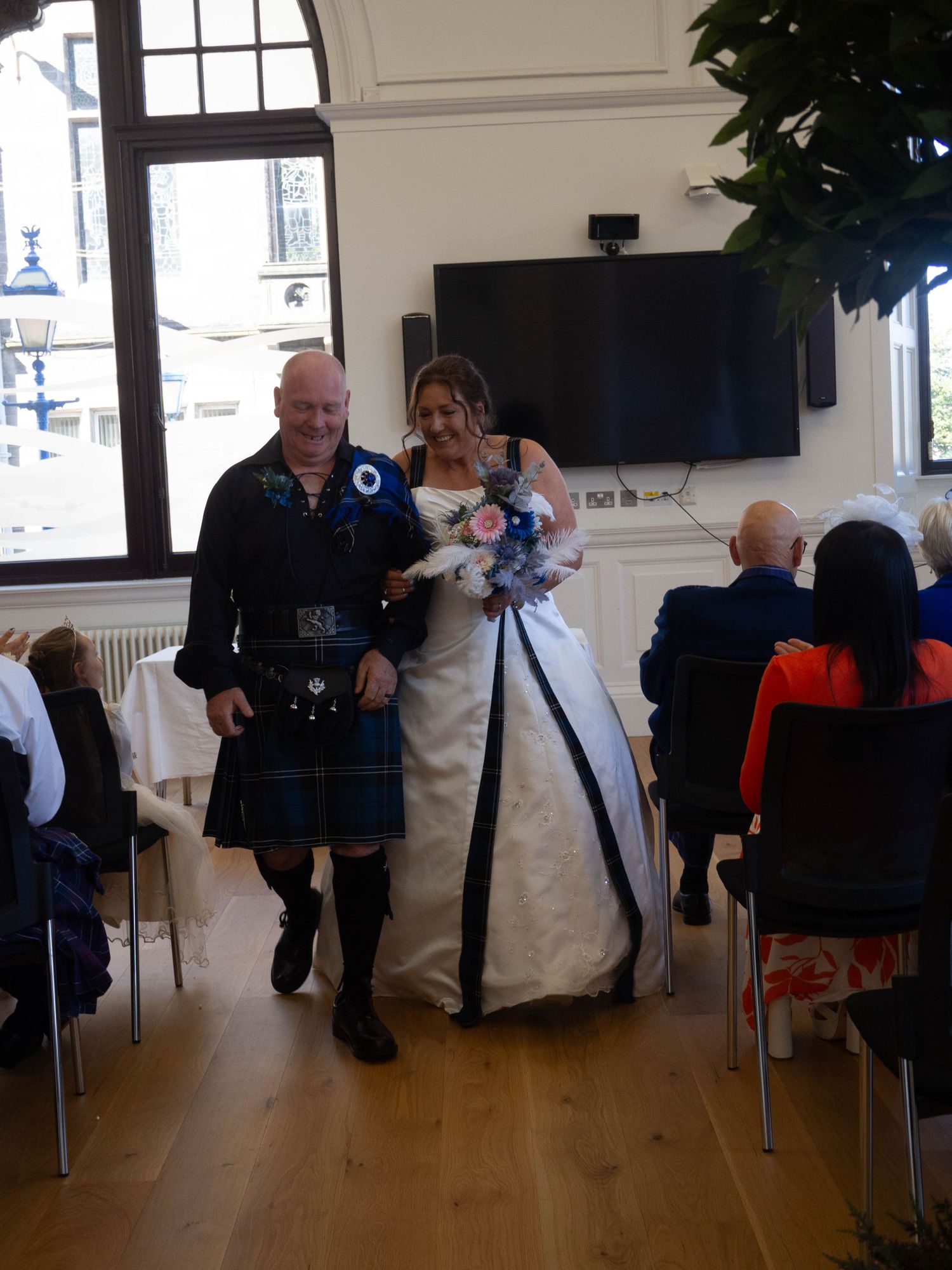 Scottish groom in kilt and bride in white and blue gown walking together at indoor wedding ceremony.