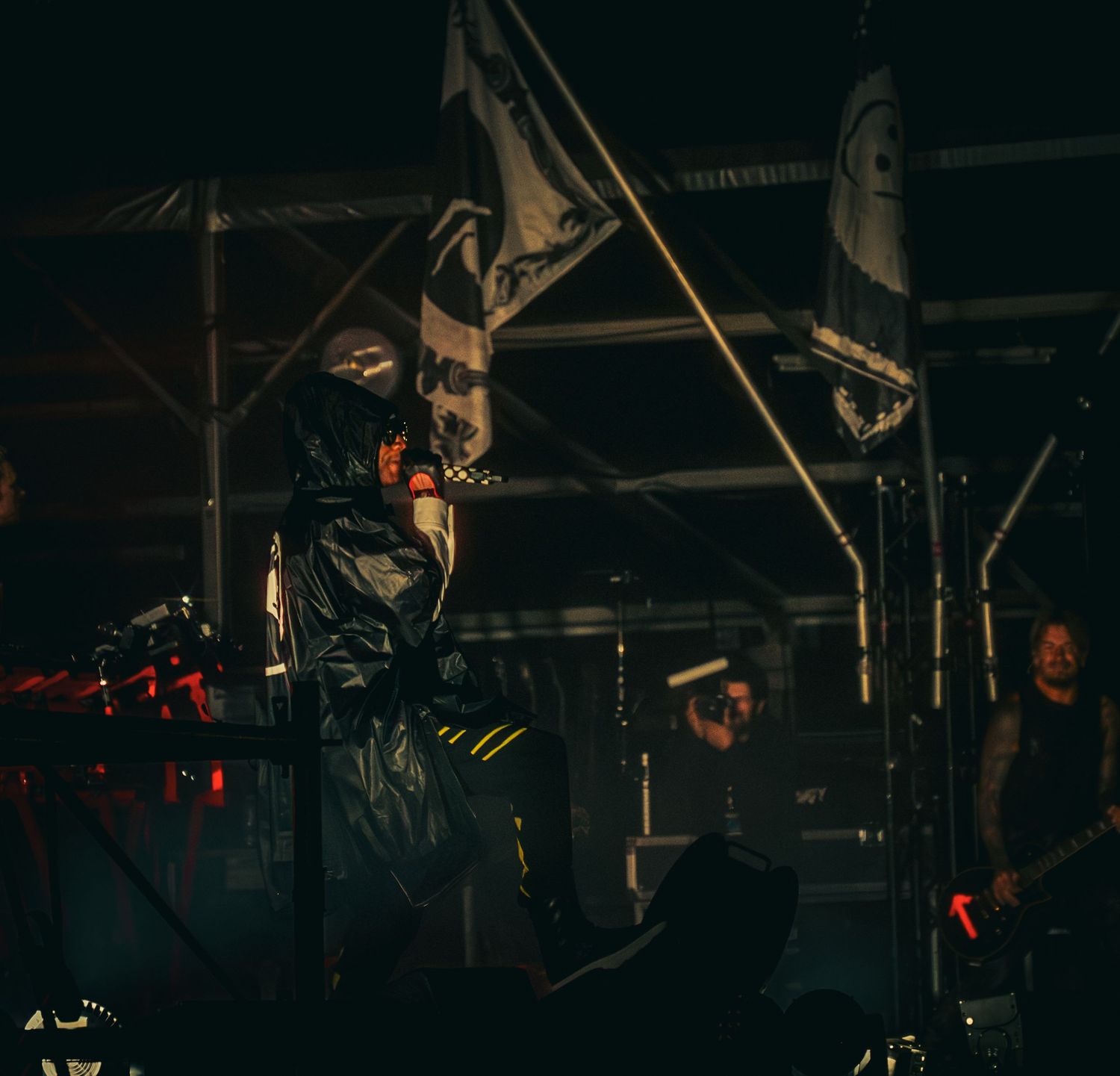 Dark concert stage with masked performer in black outfit under dramatic lighting with band members in background.