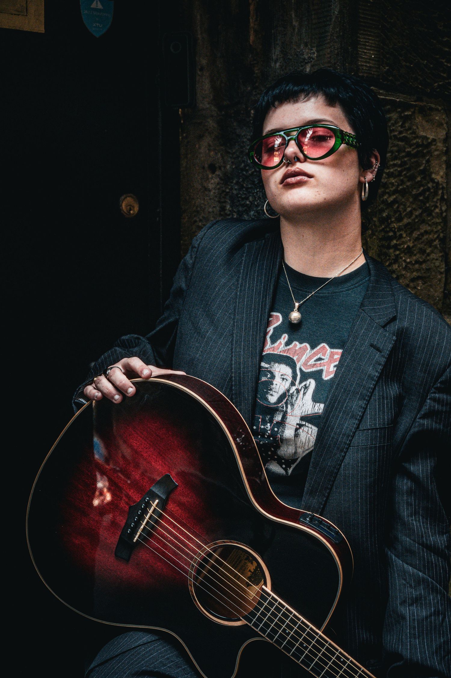 Artist holding acoustic guitar wearing red sunglasses and Prince band tee in dark alley.