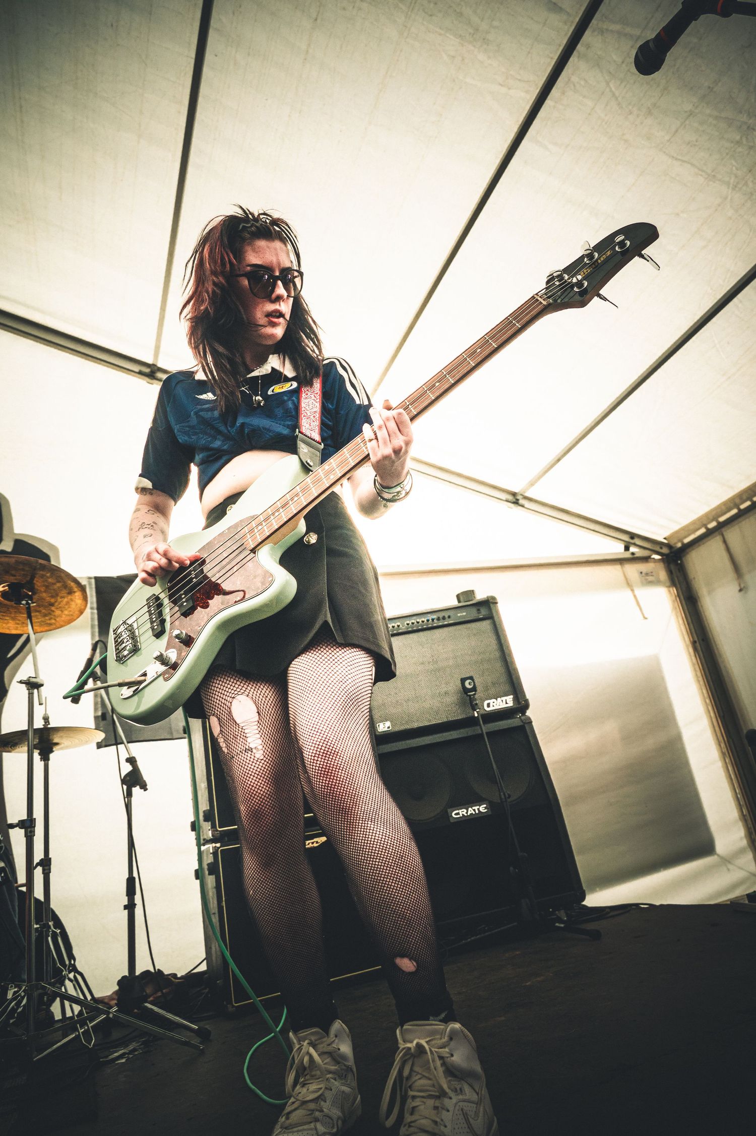Female guitarist performing on stage with a green electric guitar, wearing sunglasses and a skirt at an outdoor music festival.