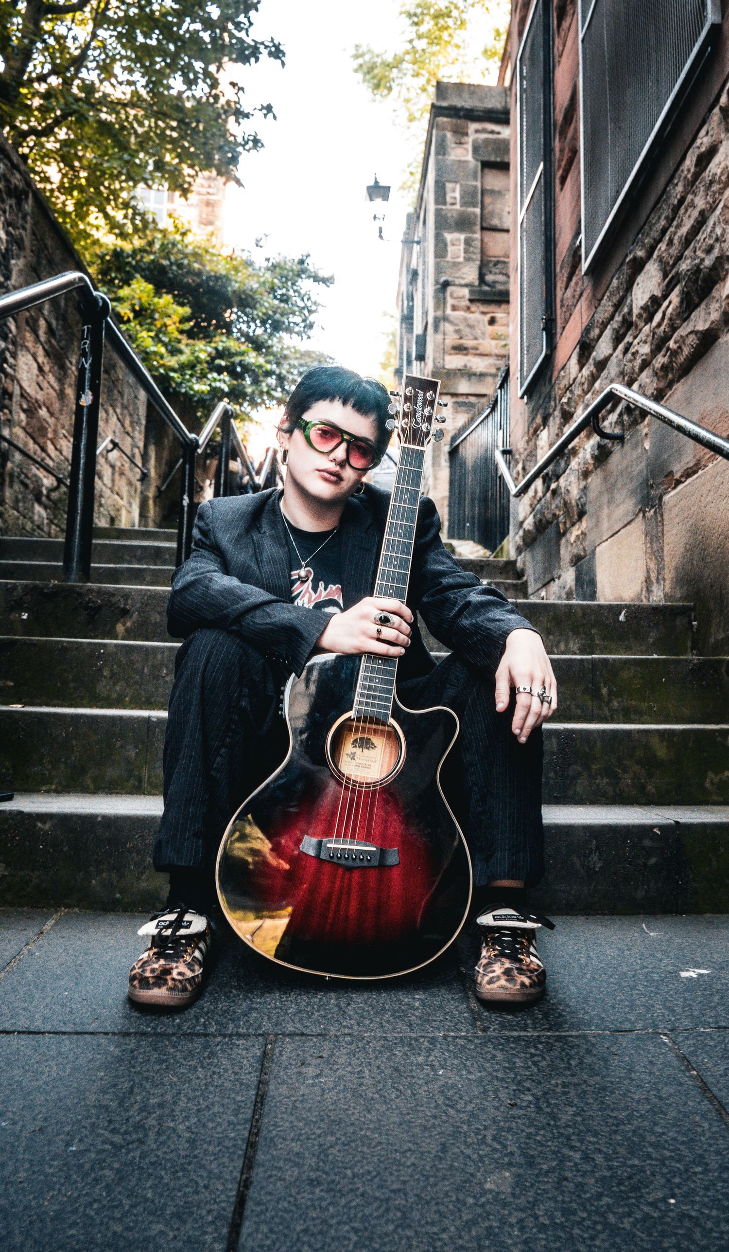 Musician sitting on urban steps holding acoustic guitar, wearing red sunglasses and black suit.