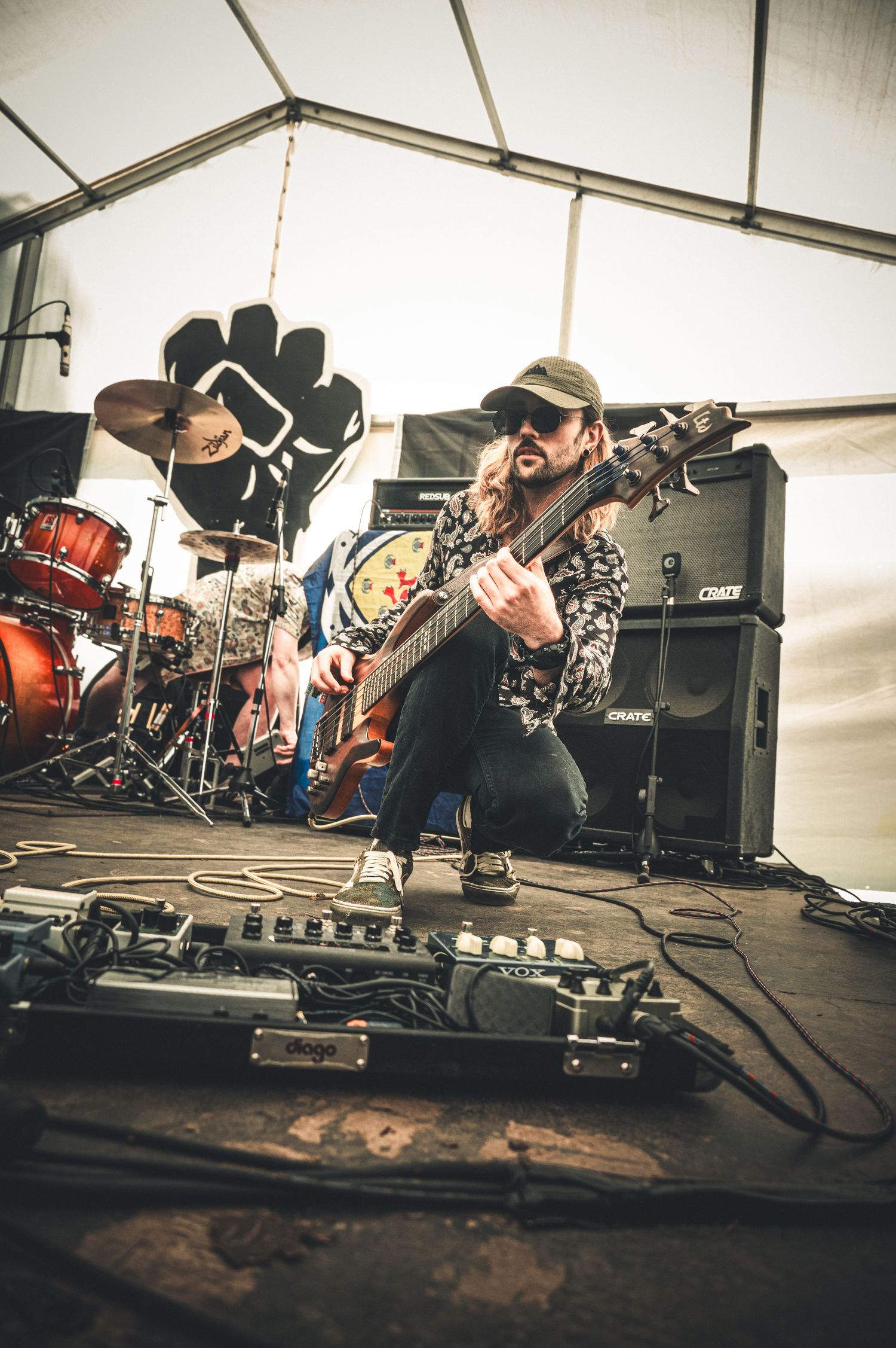 Guitarist crouching on stage with band equipment and drummer visible in background.