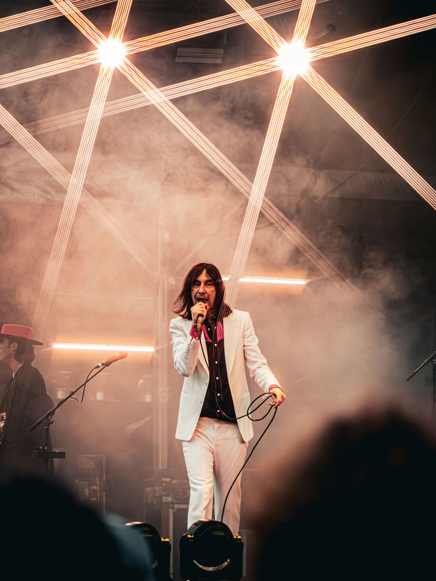 Male singer in white suit performs on smoky stage with dramatic cross lighting at live concert.