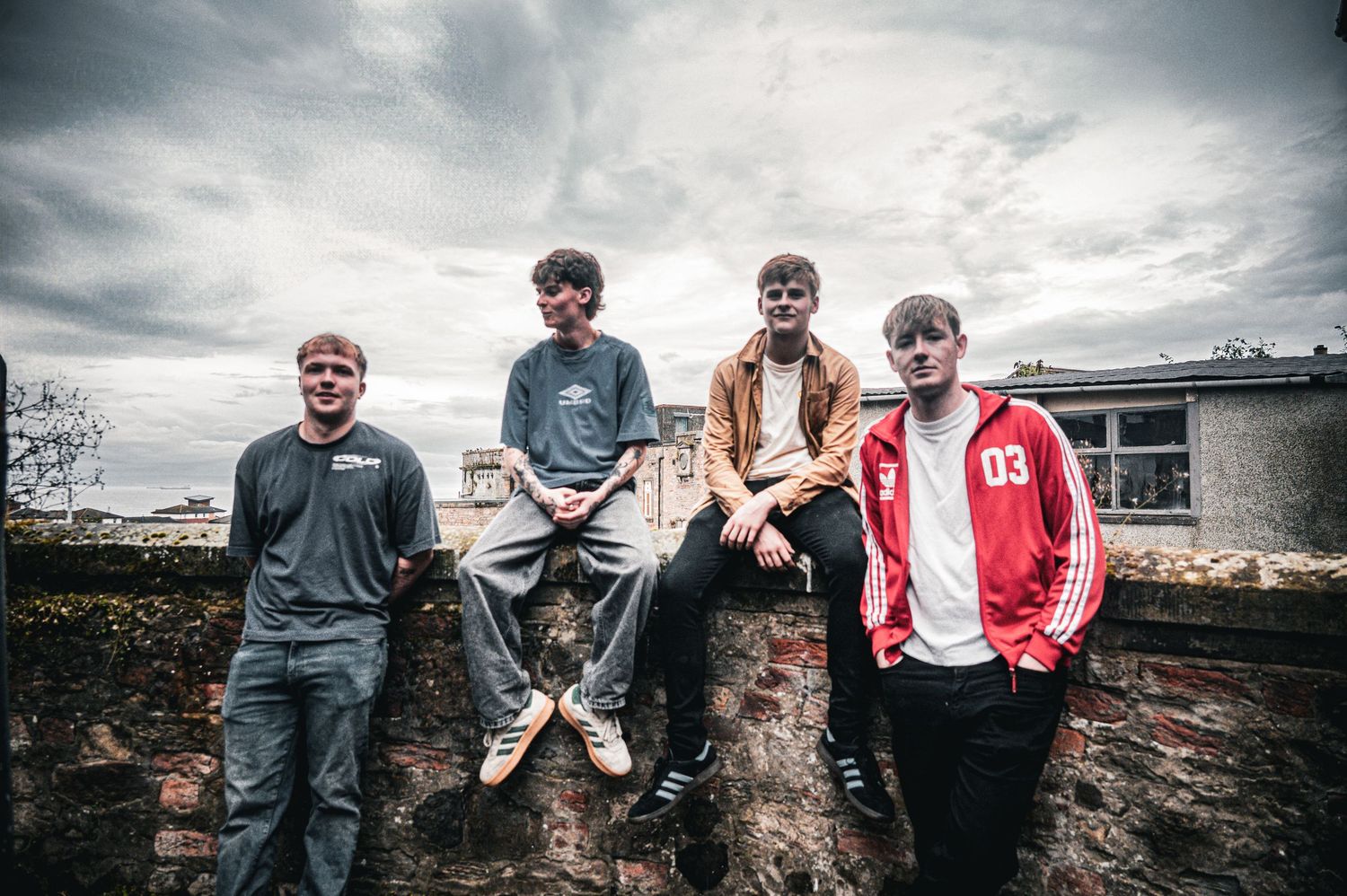 Four young men sit and stand on a stone wall outdoors under a cloudy sky.