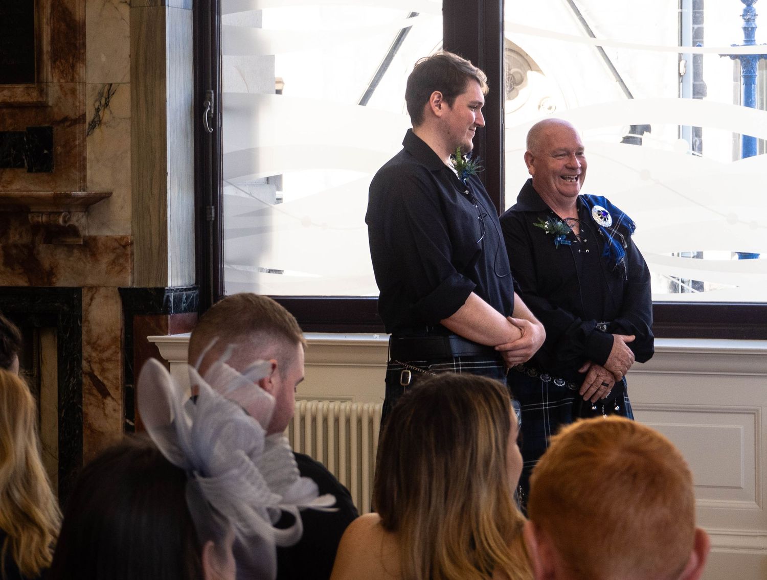 Two men in kilts standing at a Scottish wedding ceremony, one wearing a blue flower corsage, in an elegant venue.