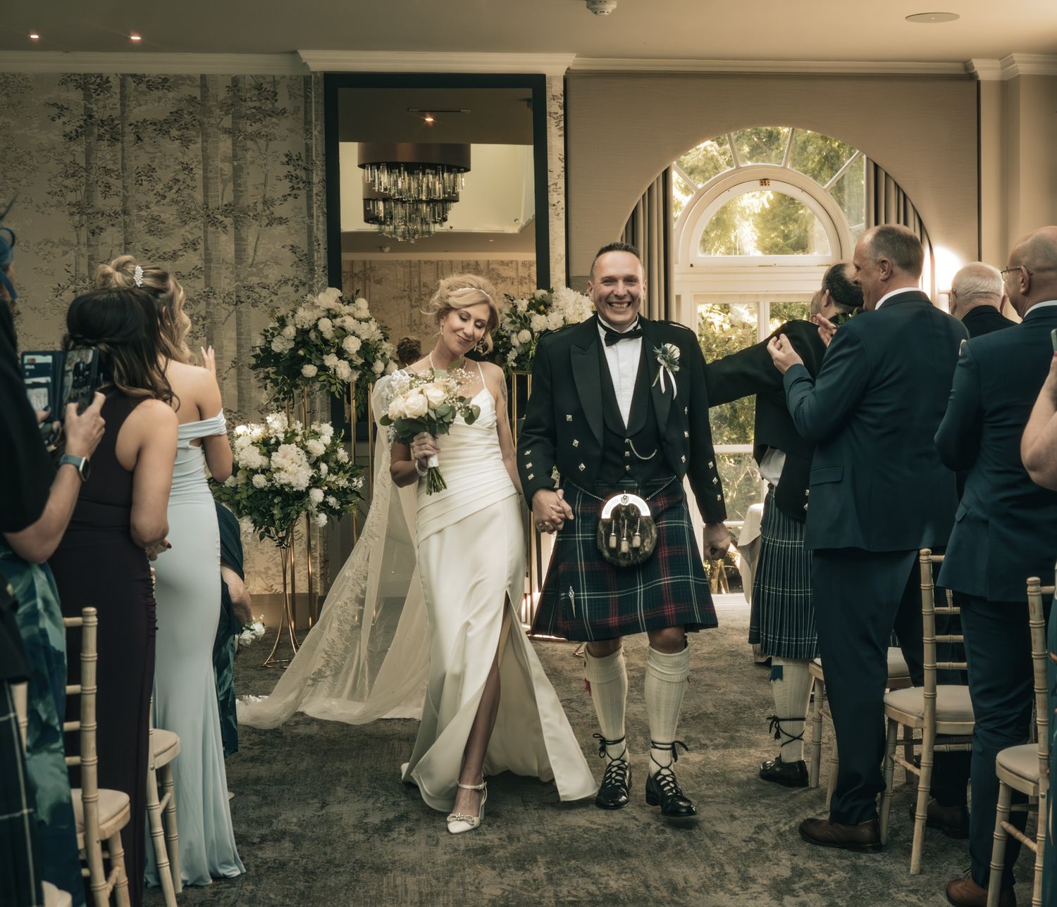 Newlyweds walk down aisle, bride in white gown, groom in tartan kilt, guests applauding at indoor ceremony.