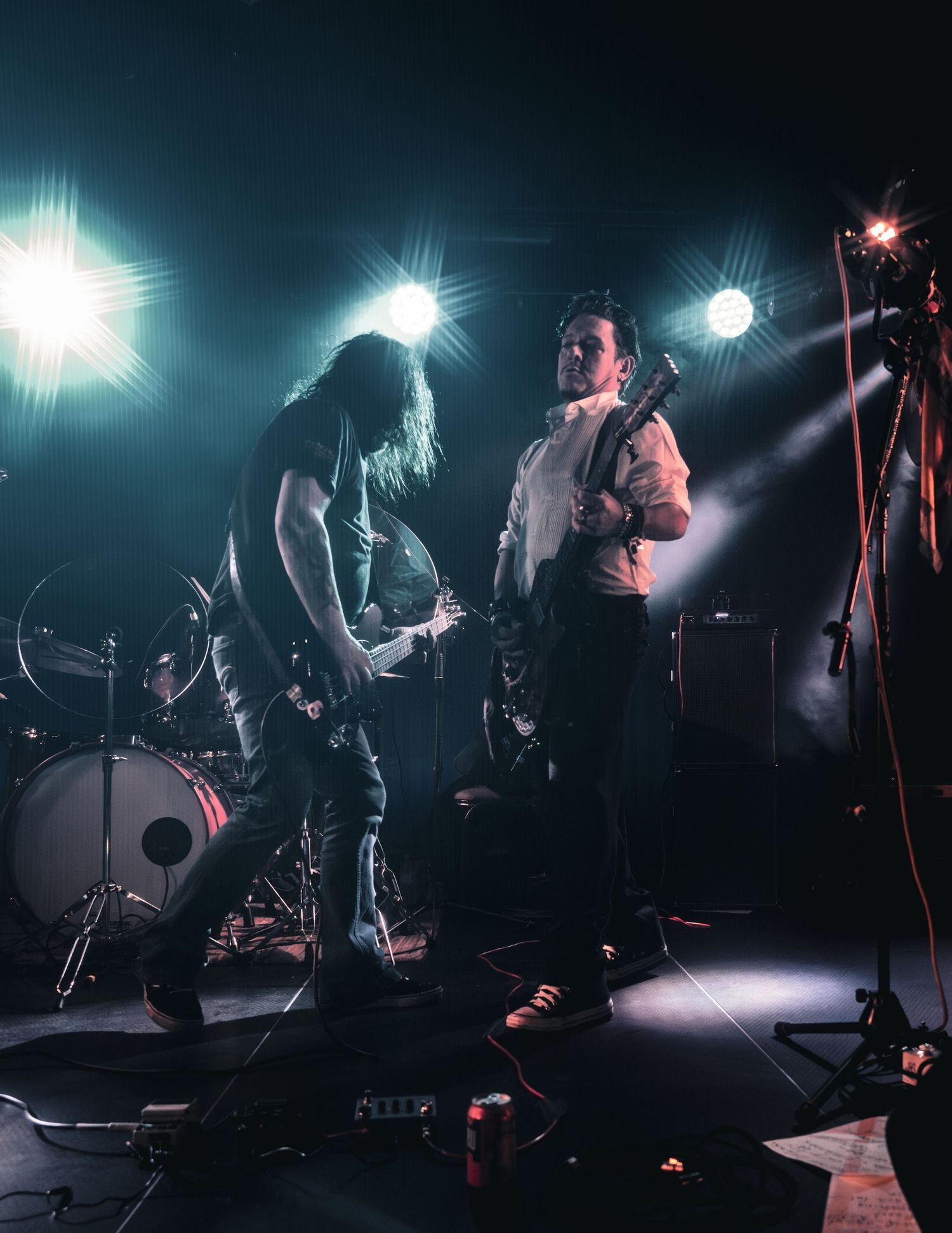 Two rock musicians perform on a dimly lit stage with dramatic spotlights, one playing guitar while a drummer is visible in the background.