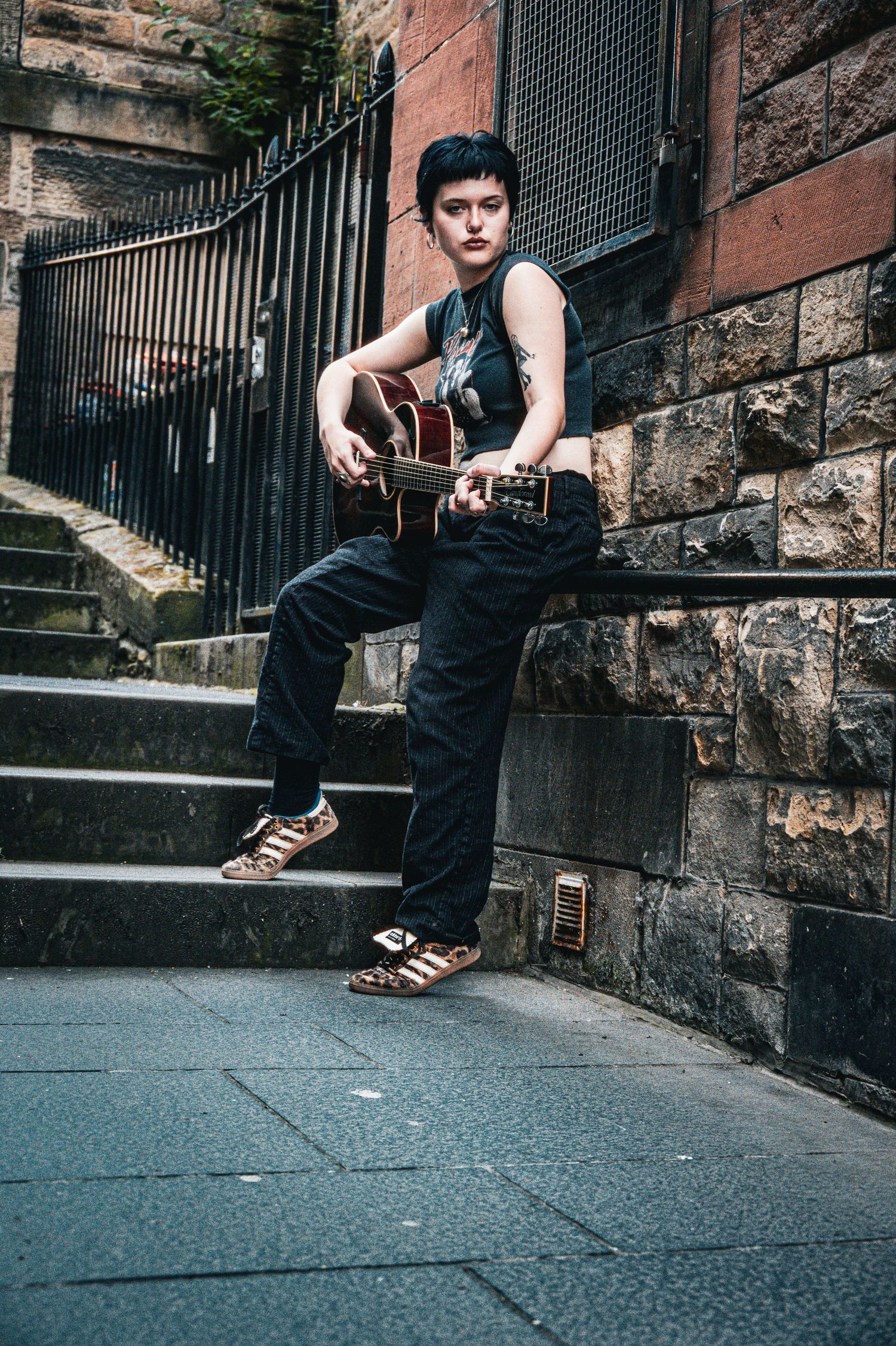 Female guitarist playing acoustic guitar while leaning against stone wall on urban stairway.