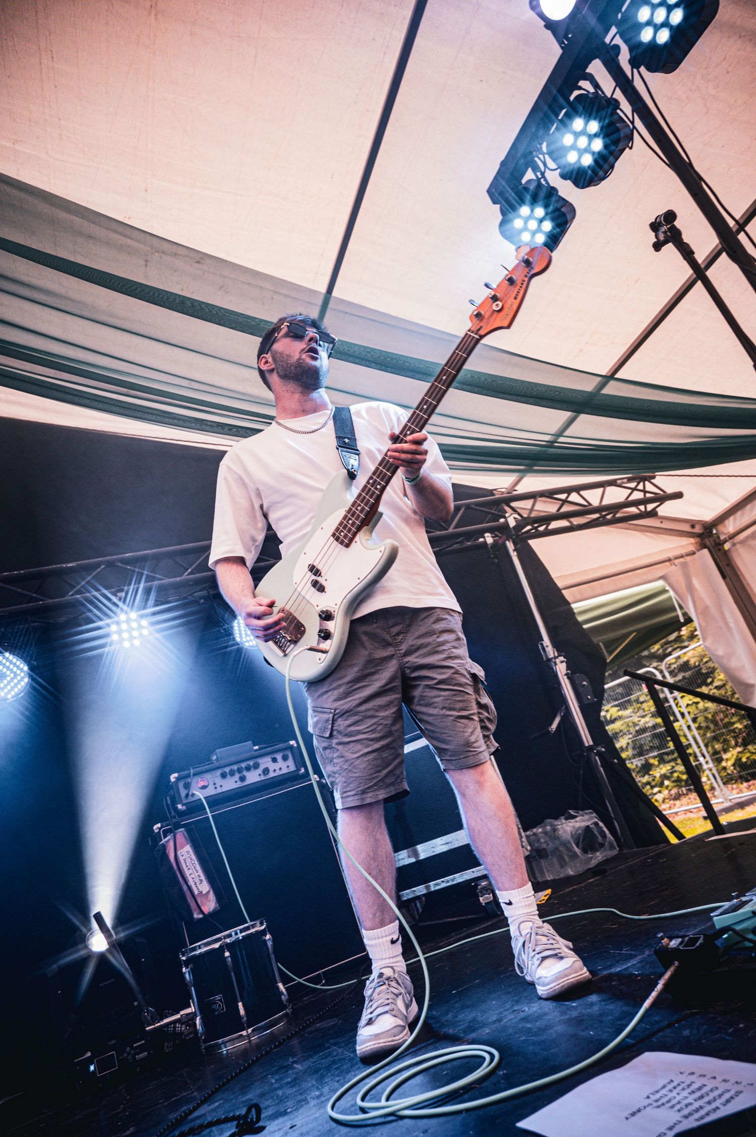 Bass guitarist performing on stage in white tee and shorts under colorful stage lights at outdoor tent venue.